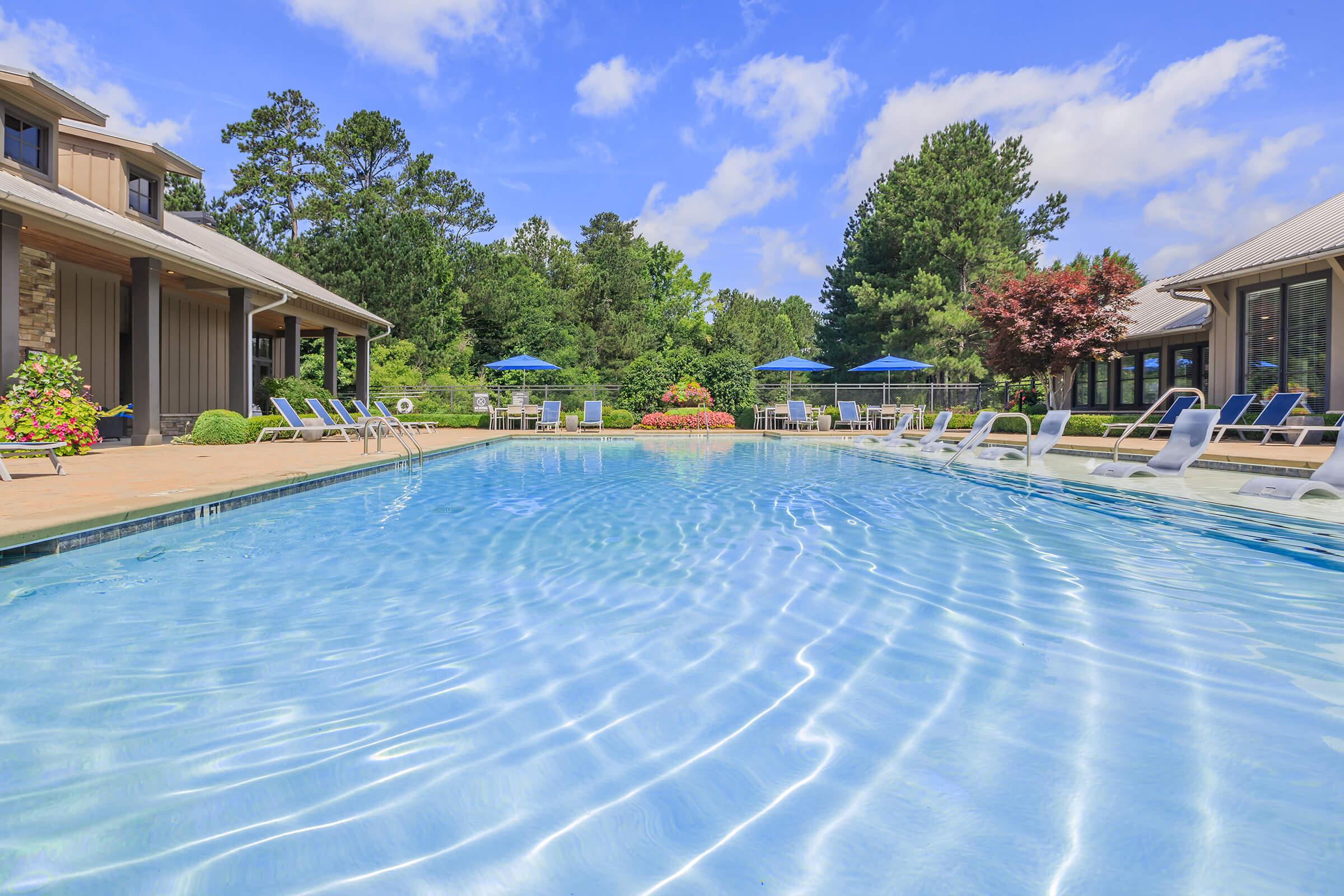 A serene swimming pool surrounded by lush greenery and colorful flowerbeds. Sun loungers are positioned along the poolside, and blue umbrellas provide shade. The clear blue water reflects the bright sky and vibrant surroundings, creating a relaxing outdoor atmosphere.