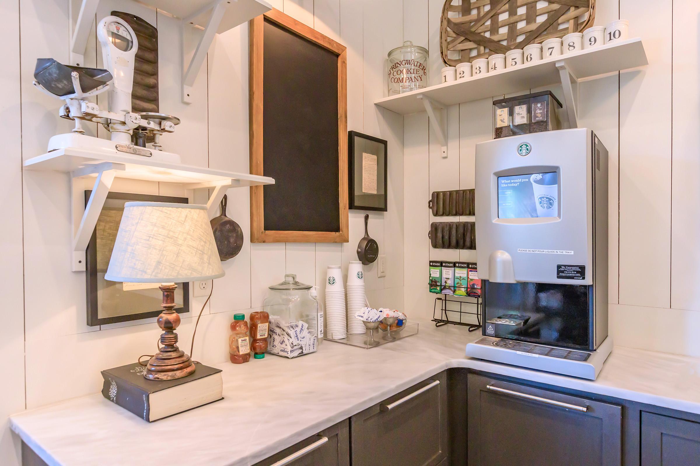 A cozy kitchen corner featuring a Starbucks coffee machine, a lamp on a stacked book, and various coffee condiments. Shelves hold a chalkboard, decorative items, and beverage supplies, creating an inviting coffee station atmosphere.