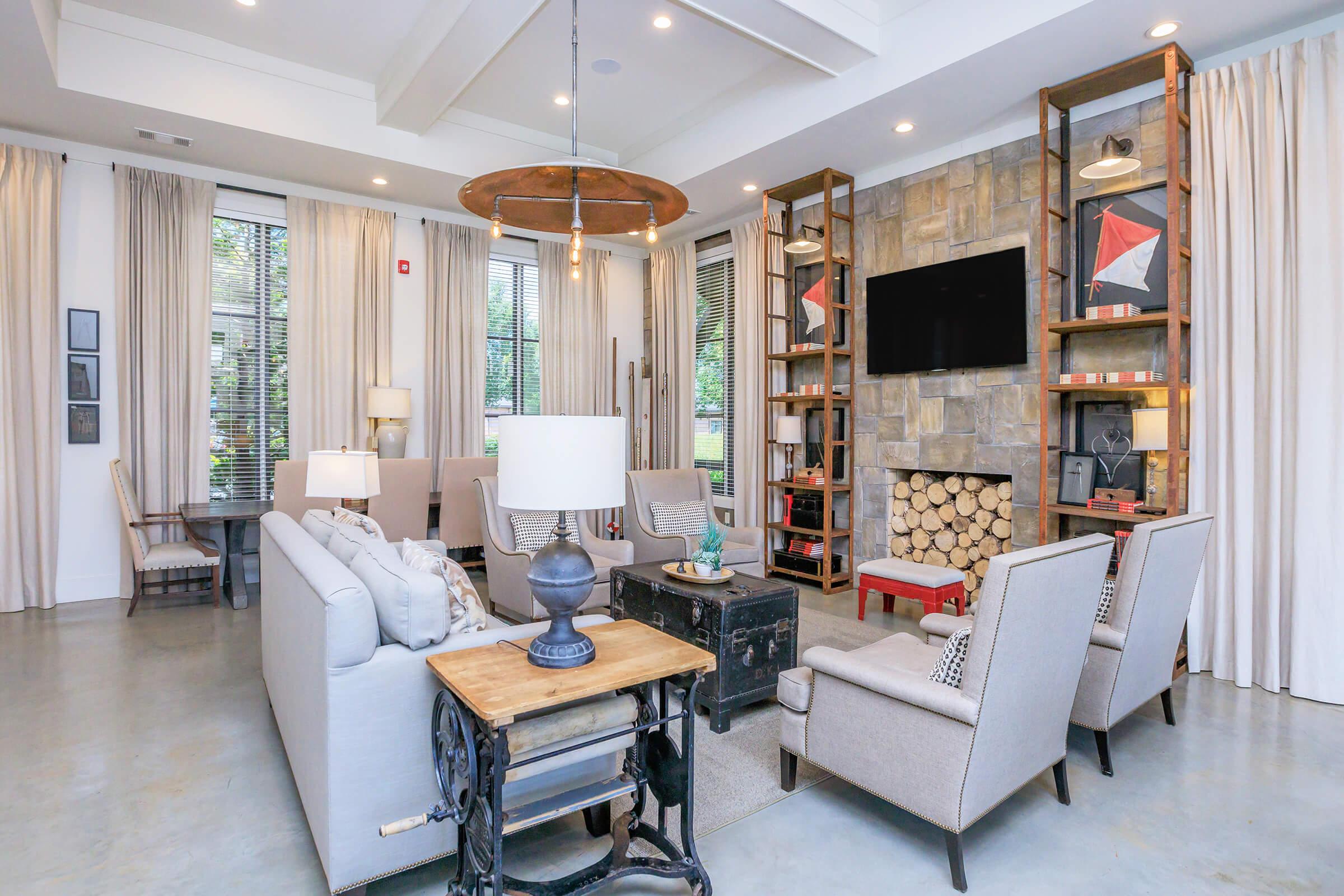 A stylish living area featuring comfortable seating with gray armchairs and a sofa. A stone fireplace with stacked logs, a large flat-screen TV mounted on the wall, and modern decor elements. Large windows with sheer curtains allow natural light, highlighting the contemporary design and tranquil atmosphere.