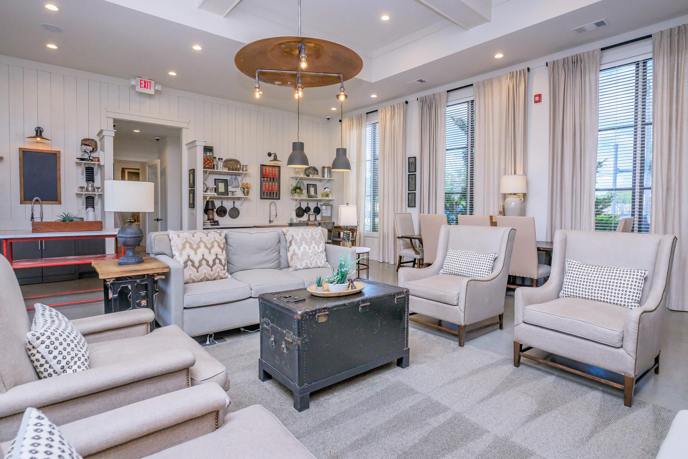 A cozy, well-lit living room featuring a gray sofa and light-colored armchairs arranged around a dark coffee table. Soft, neutral decor with curtains, decorative items on shelves, and warm lighting create an inviting atmosphere. The space is stylishly furnished with a modern touch.