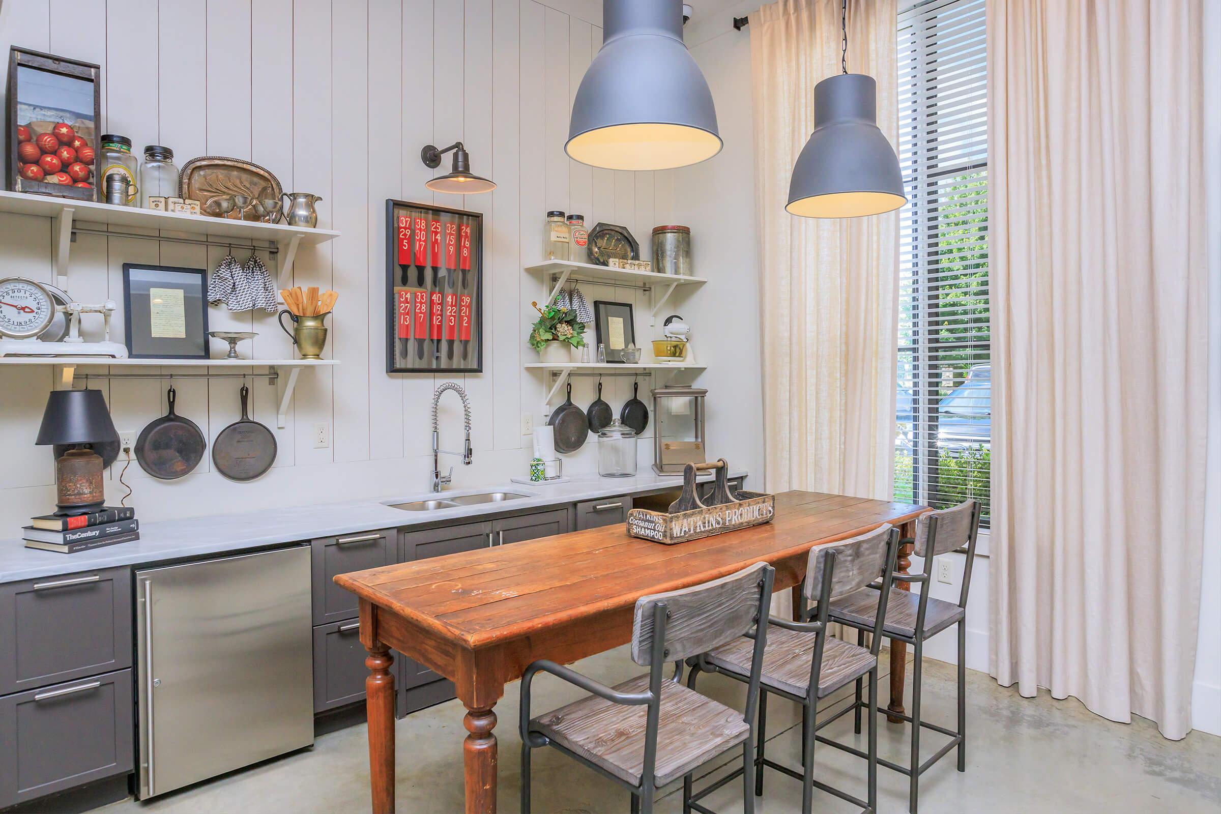 A modern kitchen featuring a wooden dining table with three chairs, grey cabinetry, and open shelves displaying cookware and decorative items. Large windows with curtains provide natural light, and pendant lights hang overhead, creating a stylish and functional space.