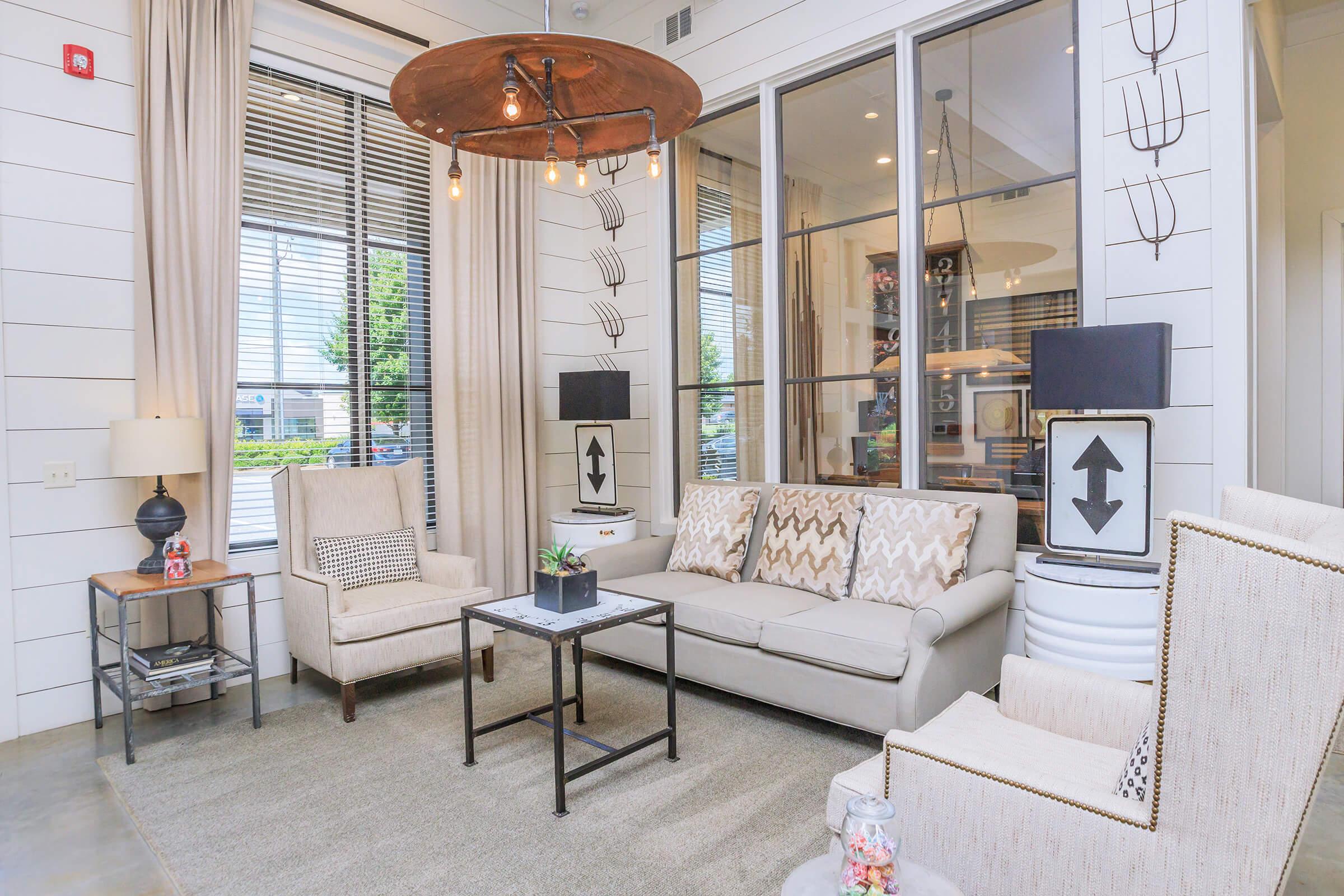 A cozy living room with large windows letting in natural light. It features a light-colored sofa with patterned cushions, two armchairs, a small coffee table, and decorative lamps. The walls are white shiplap, and there are curtains framing the windows, creating a warm and inviting atmosphere.