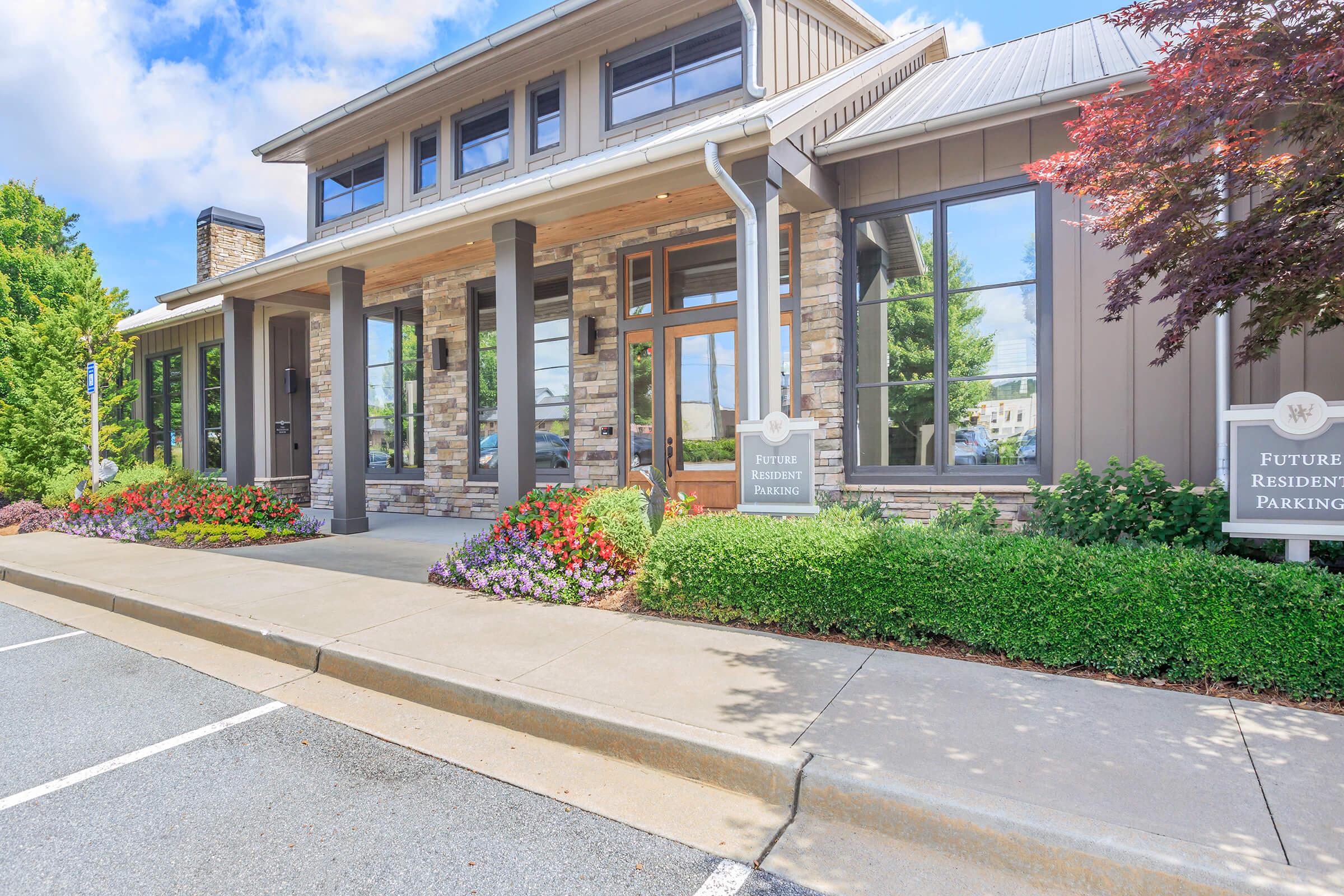 Exterior view of a modern building with a large entrance featuring glass doors and stone accents. Colorful flower beds line the walkway, and a sign indicates "Future Residents Parking." The surrounding area includes well-maintained landscaping and blue skies.