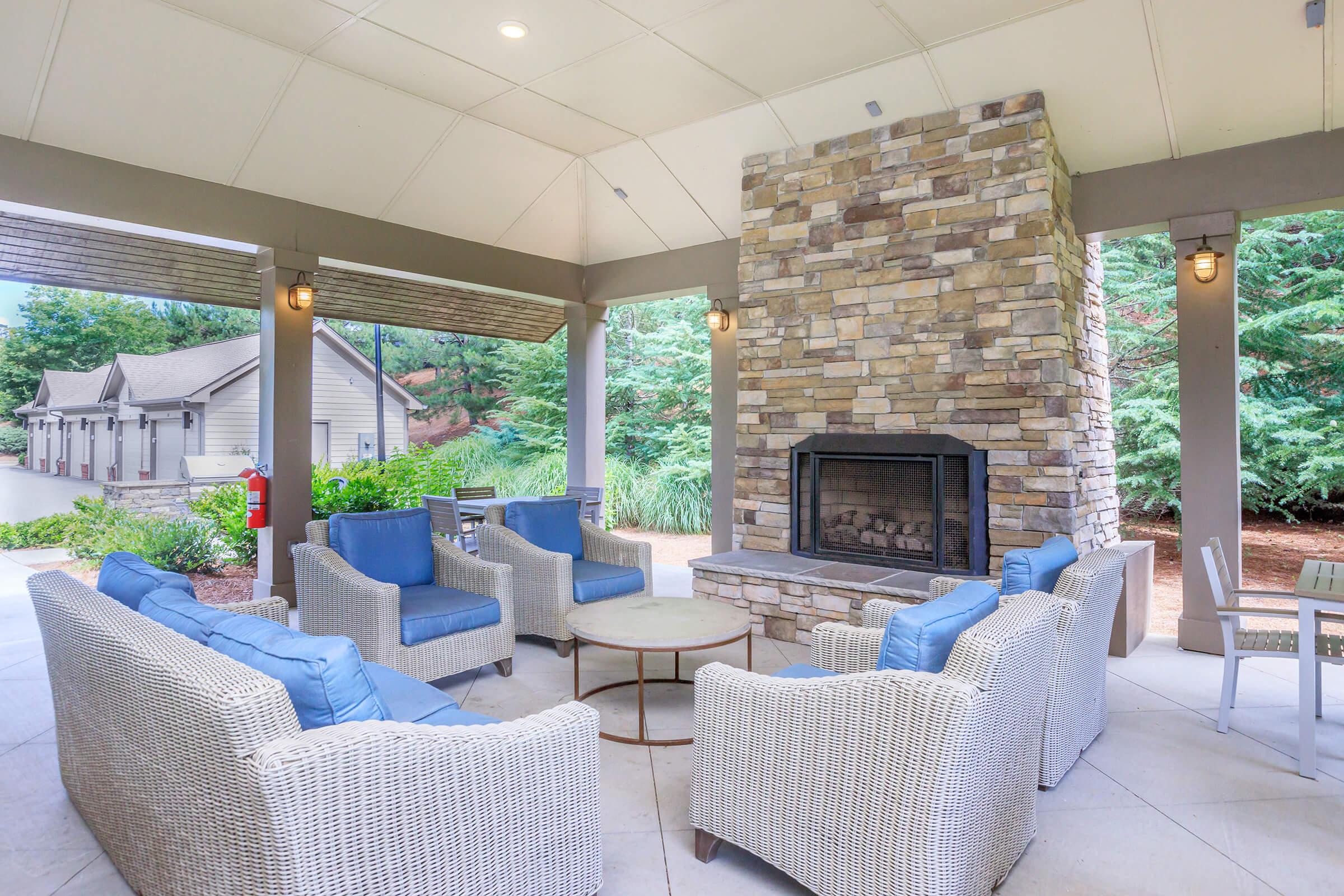 A cozy outdoor lounge area featuring a stone fireplace, surrounded by comfortable seating with blue cushions. The space is under a covered patio, with greenery visible in the background, creating a relaxing atmosphere for gatherings or casual conversations.