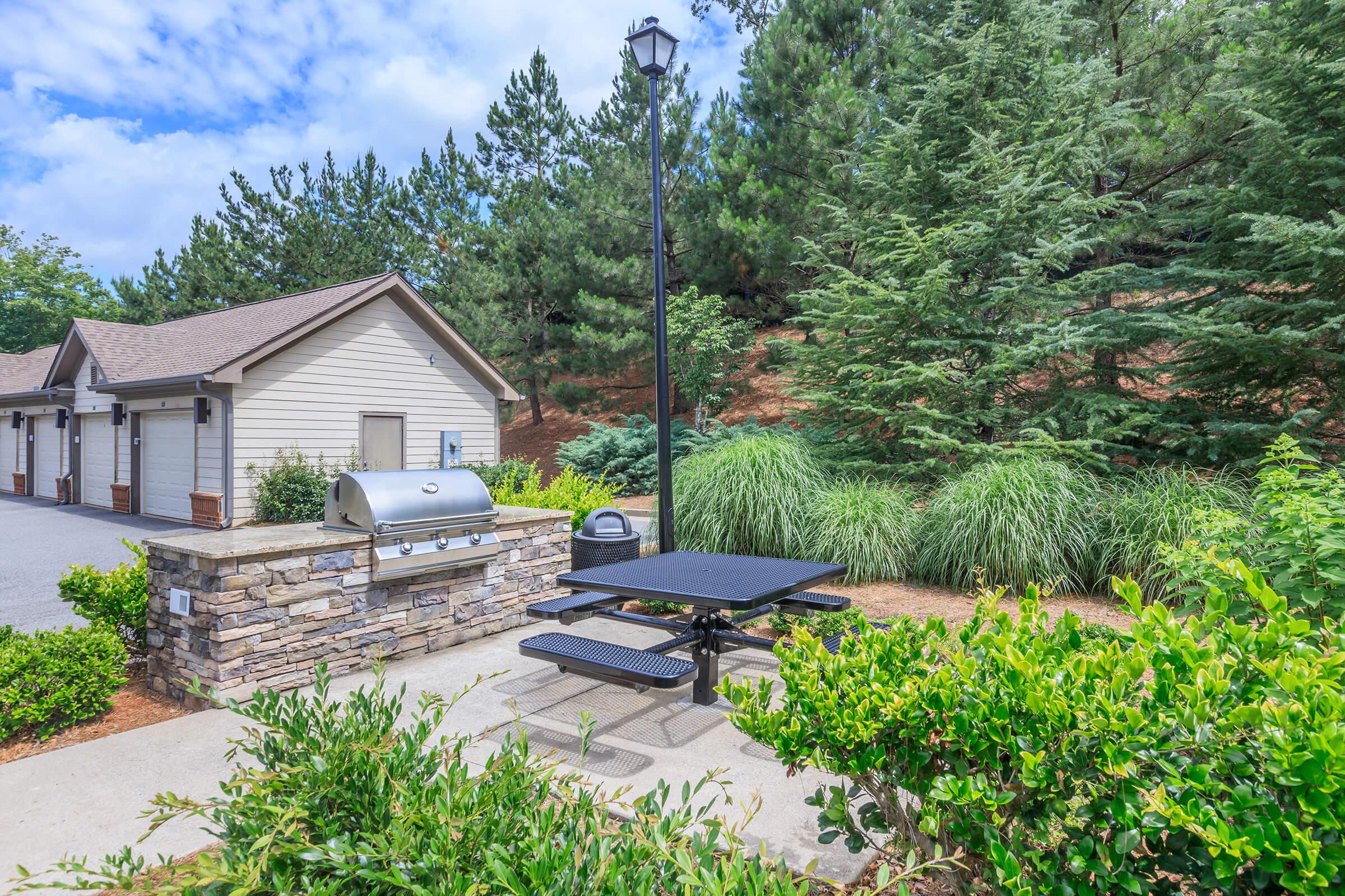 A clean outdoor space featuring a picnic table next to a stone grilling area. In the background, a well-maintained building and lush greenery, including trees and shrubs, create a serene atmosphere. A lamp post adds to the ambiance, making it a nice spot for gatherings or relaxation.