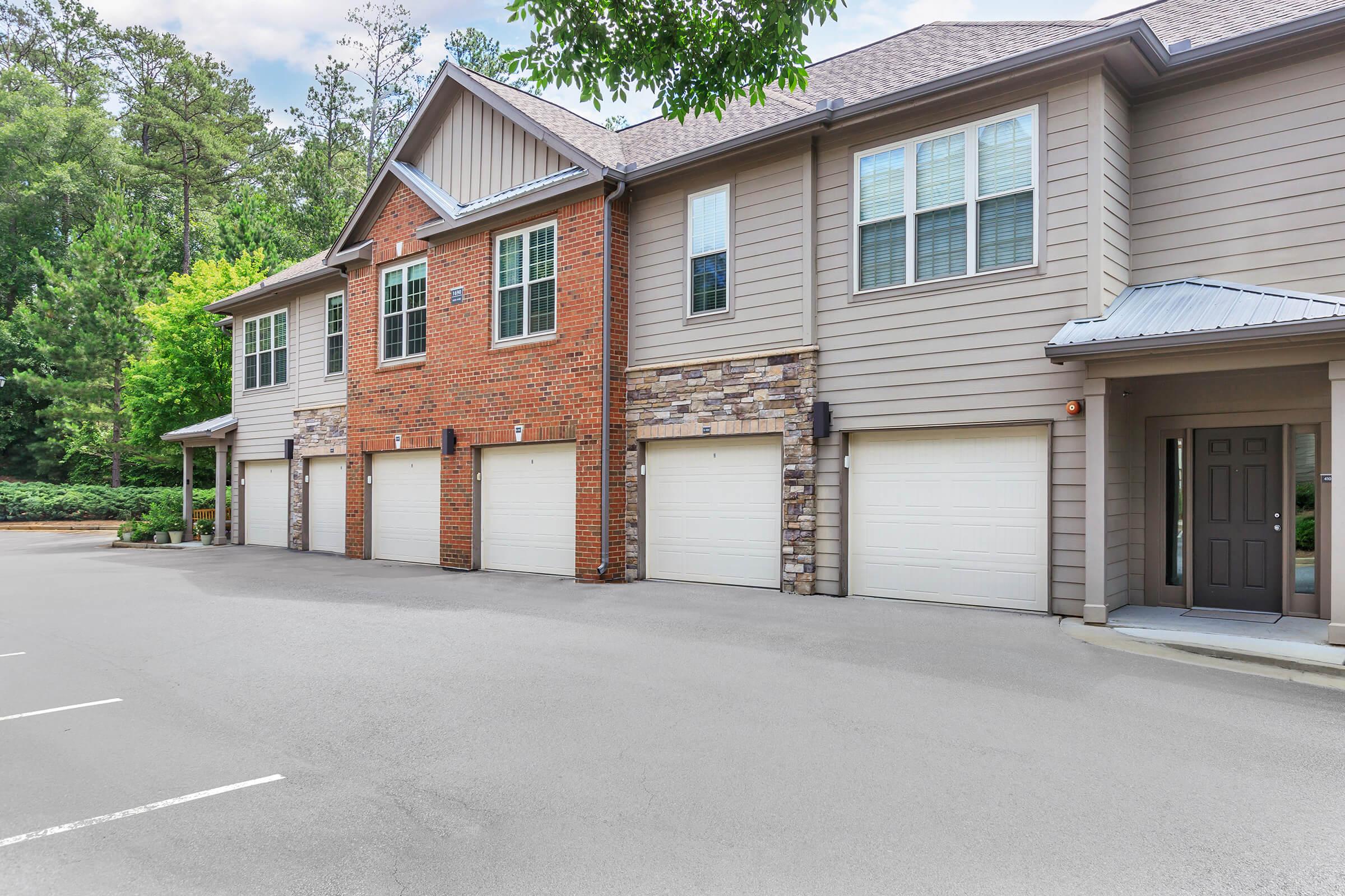 A row of attached townhouses featuring a mix of brick and siding exteriors. The garages are positioned in front of each unit, with a driveway leading to them. Surrounding greenery adds a natural element to the scene, and the sky is partly cloudy, suggesting a pleasant day.