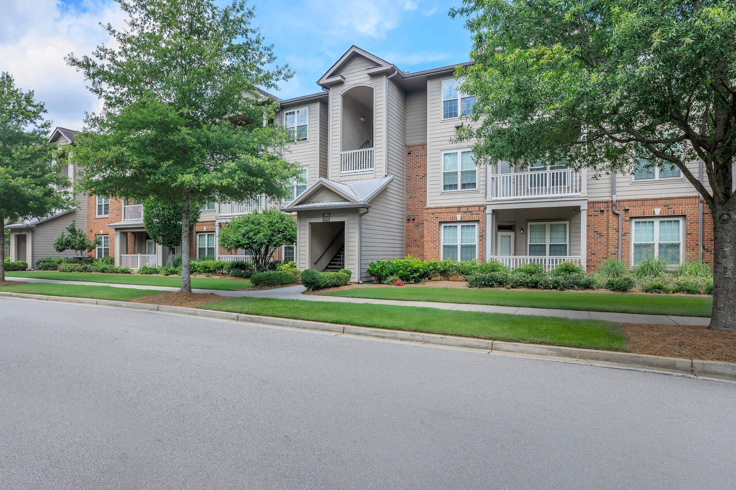 A well-maintained apartment complex featuring three-story buildings with a mix of brick and siding. The landscaped front yard includes trees and shrubs, and a paved walkway leads to the entrance. The scene captures a sunny day with a clear sky, showcasing the inviting atmosphere of the community.