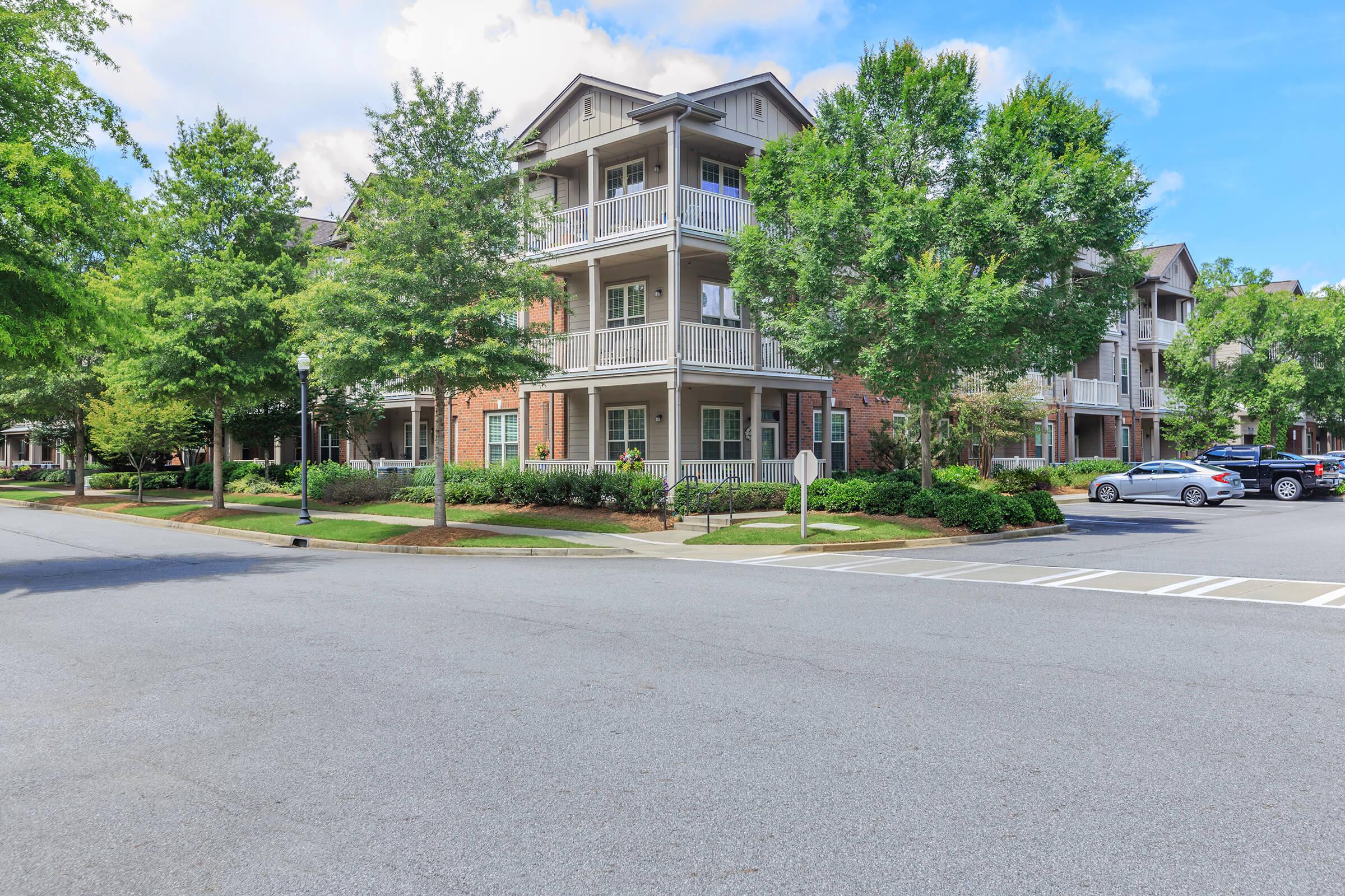 A residential street view featuring multi-story apartment buildings surrounded by well-maintained landscaping, including trees and bushes. There are parked cars along the road and a clear blue sky overhead, creating a peaceful suburban atmosphere.
