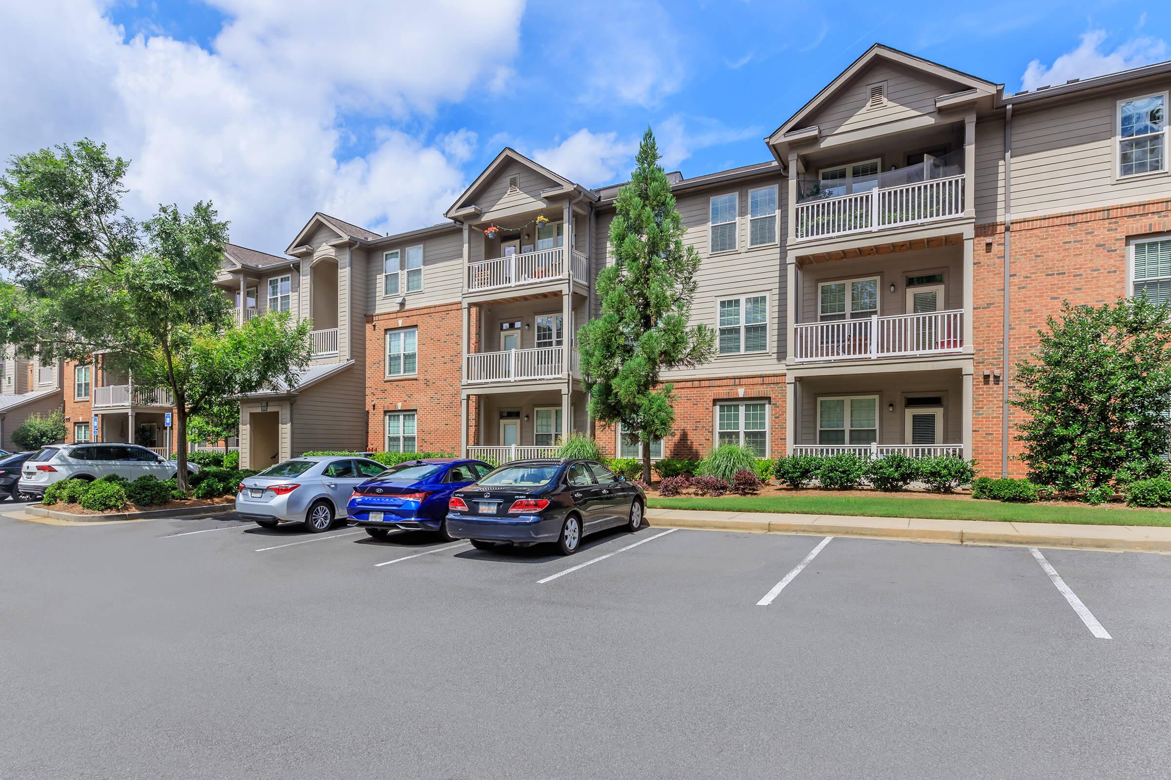 A modern multi-story apartment building featuring a mix of brick and siding exterior, with balconies on the upper floors. The surrounding area includes well-maintained landscaping and a parking lot with various cars. The sky is blue with a few clouds, suggesting a pleasant day.