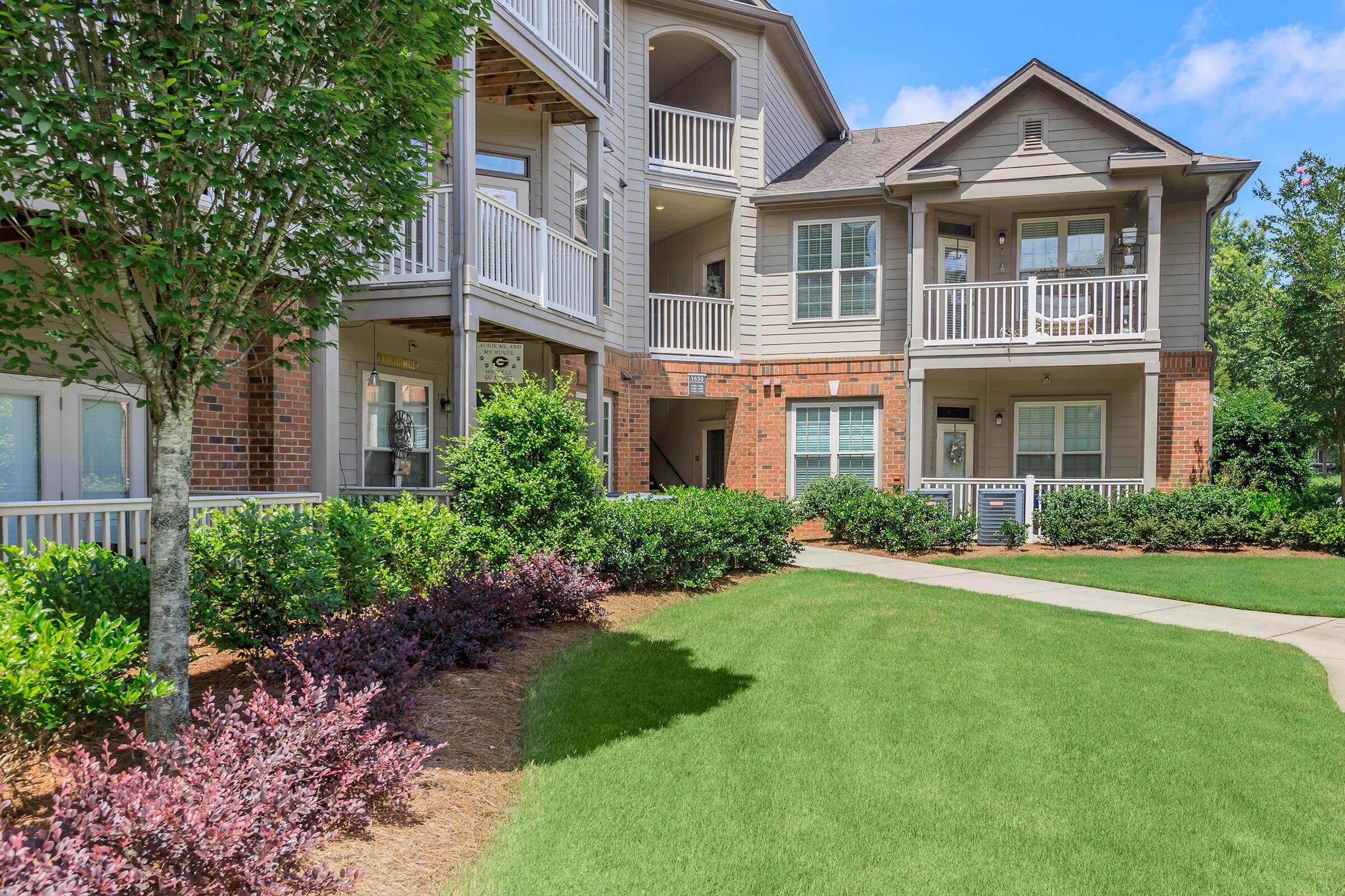 A well-maintained apartment complex featuring a mix of brick and wooden architecture. The building has multiple balconies, surrounded by lush green lawns and landscaped shrubs. Clear blue sky overhead enhances the inviting atmosphere of the outdoor space.