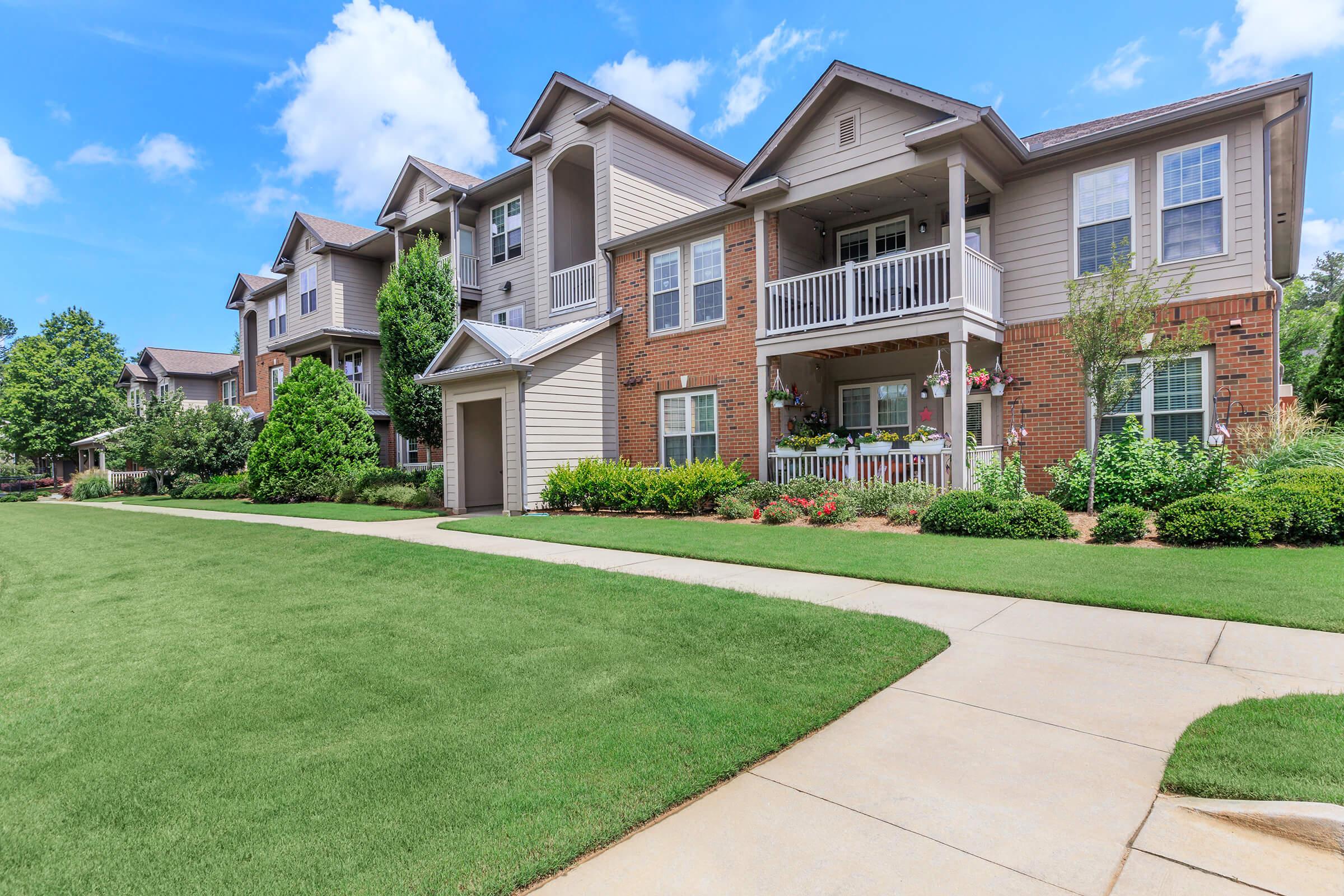 A row of modern apartment buildings with a mix of brick and light-colored siding, featuring balconies and landscaped gardens. A paved sidewalk runs alongside a lush green lawn, with trees and shrubs enhancing the exterior. The sky is partly cloudy, creating a bright and inviting atmosphere.