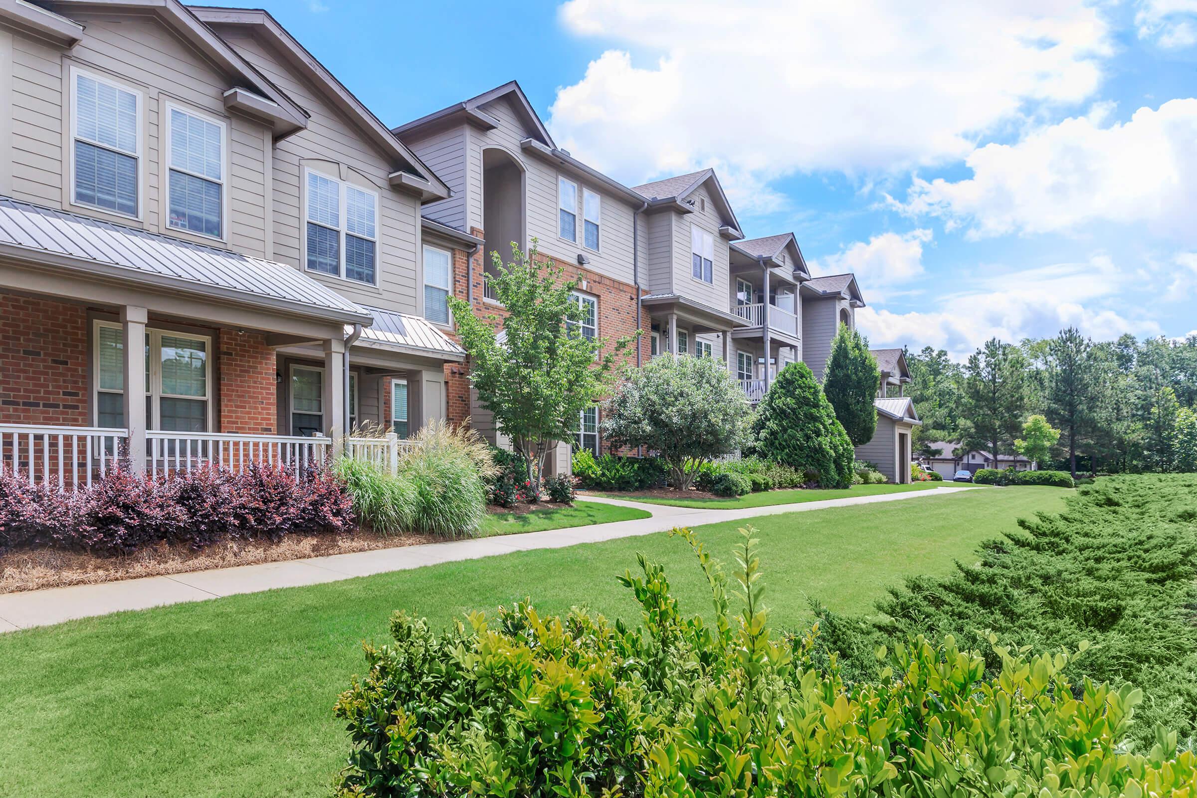 A view of a well-maintained apartment complex featuring multiple buildings with a blend of brick and siding exteriors. Neatly trimmed lawns and vibrant landscaping with shrubs and flowers enhance the outdoor space, under a bright blue sky with scattered clouds.