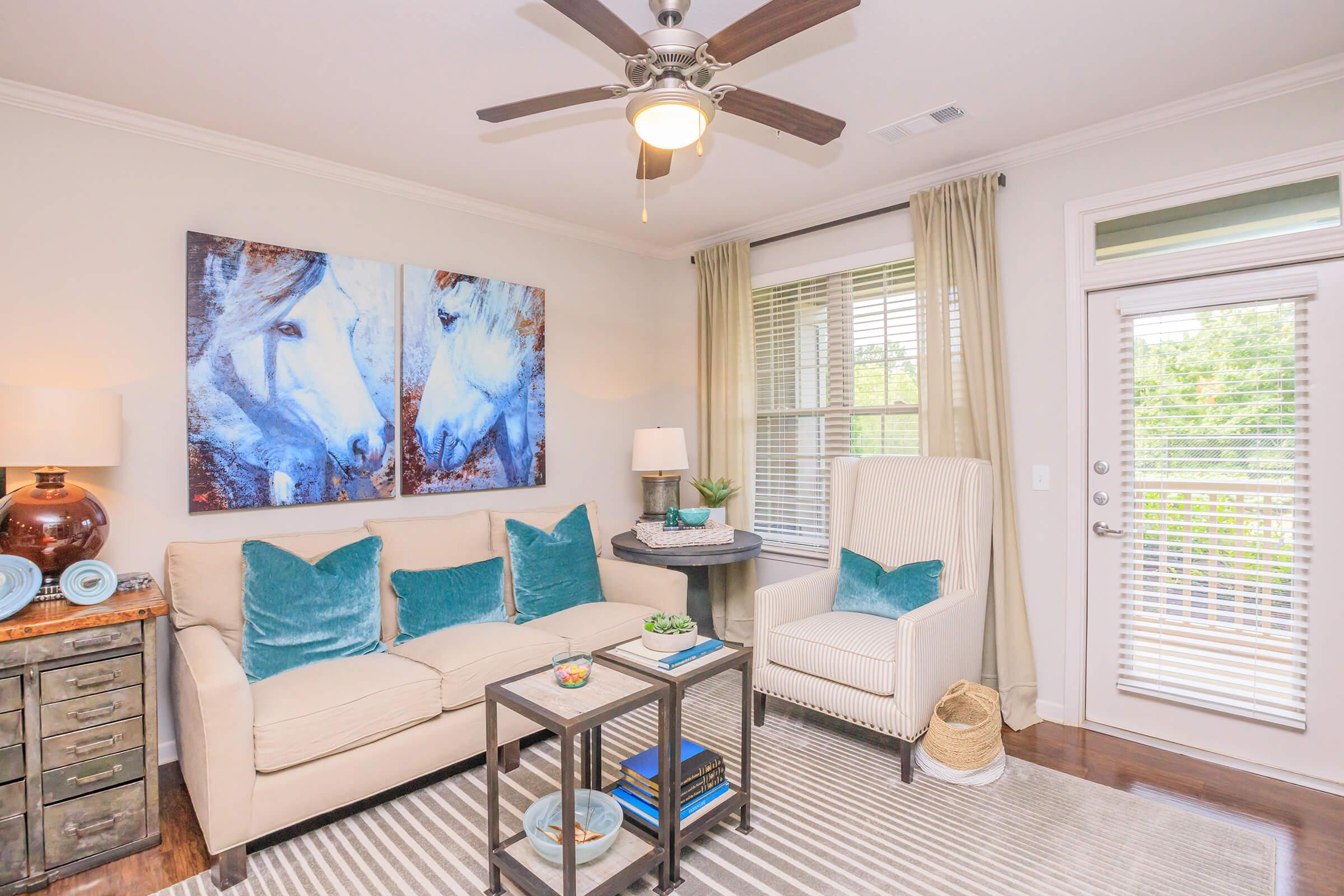 A cozy living room featuring a light beige sofa adorned with turquoise accent pillows, a small side table with decorative items, and a white armchair. The walls are decorated with a large artwork of two horses. Natural light streams in through a window with white curtains, enhancing the warm ambiance of the space.