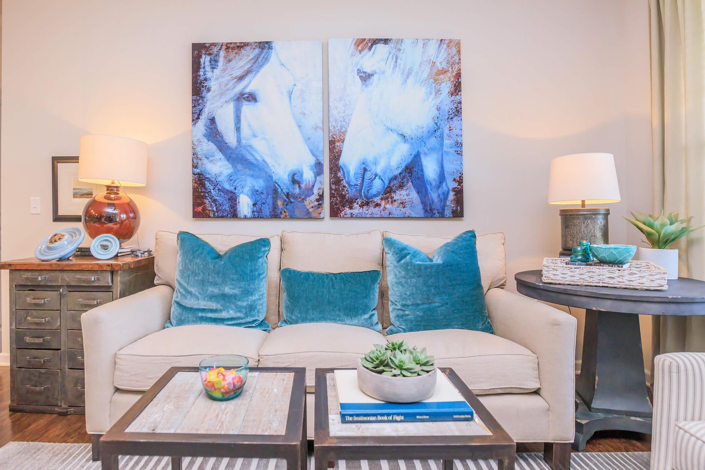 A cozy living room featuring a beige couch adorned with turquoise pillows, two large horse paintings on the wall, and a wooden coffee table. Decorative items include a bowl of colorful candies, a potted succulent, and a small table lamp. The ambiance is warm and inviting, complemented by soft lighting.