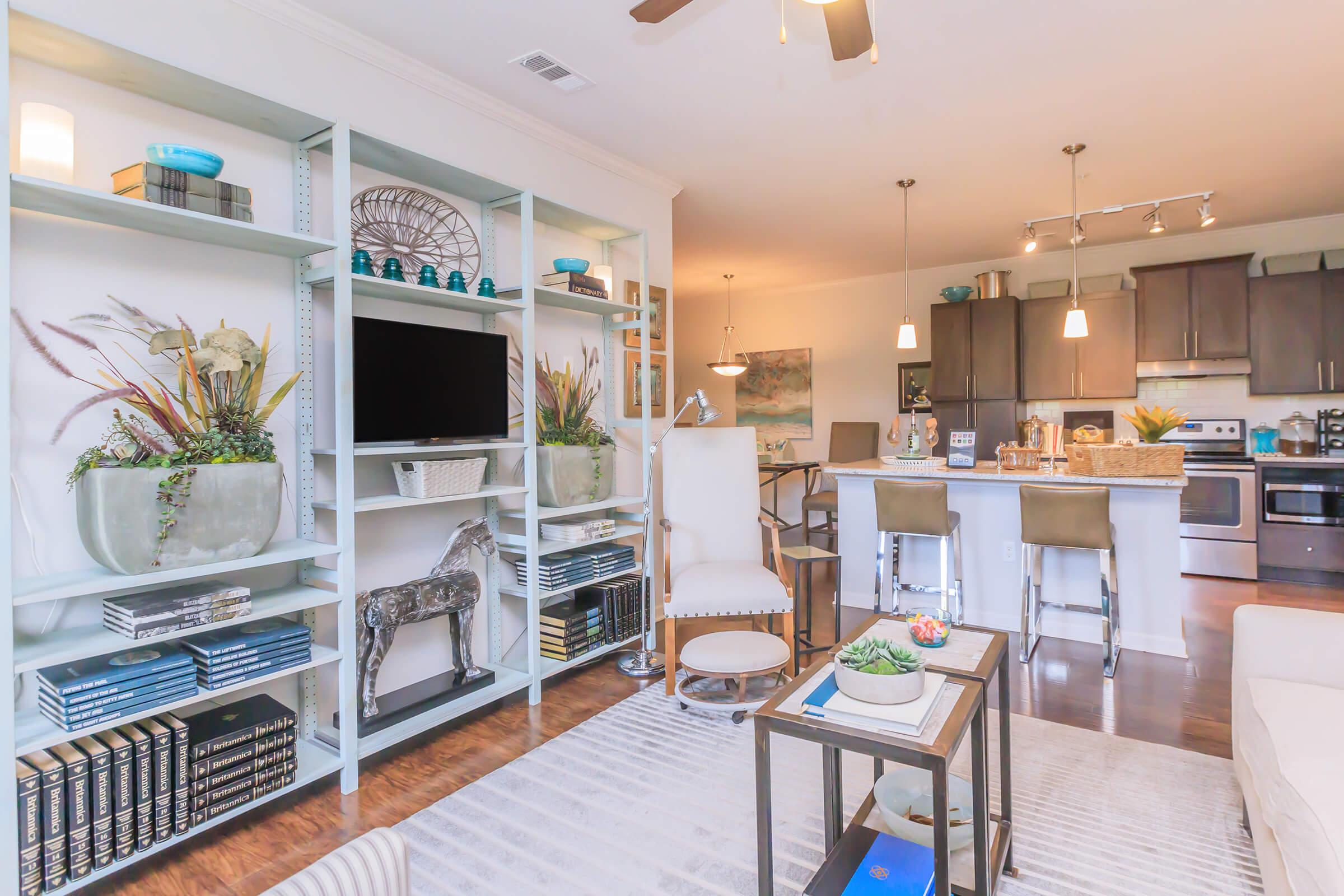Bright and modern living room featuring a light-colored sofa, a sleek coffee table, and stylish shelving with decorative plants and books. In the background, there's an open kitchen with stainless steel appliances and a dining area illuminated by pendant lights. The space is inviting and well-organized.