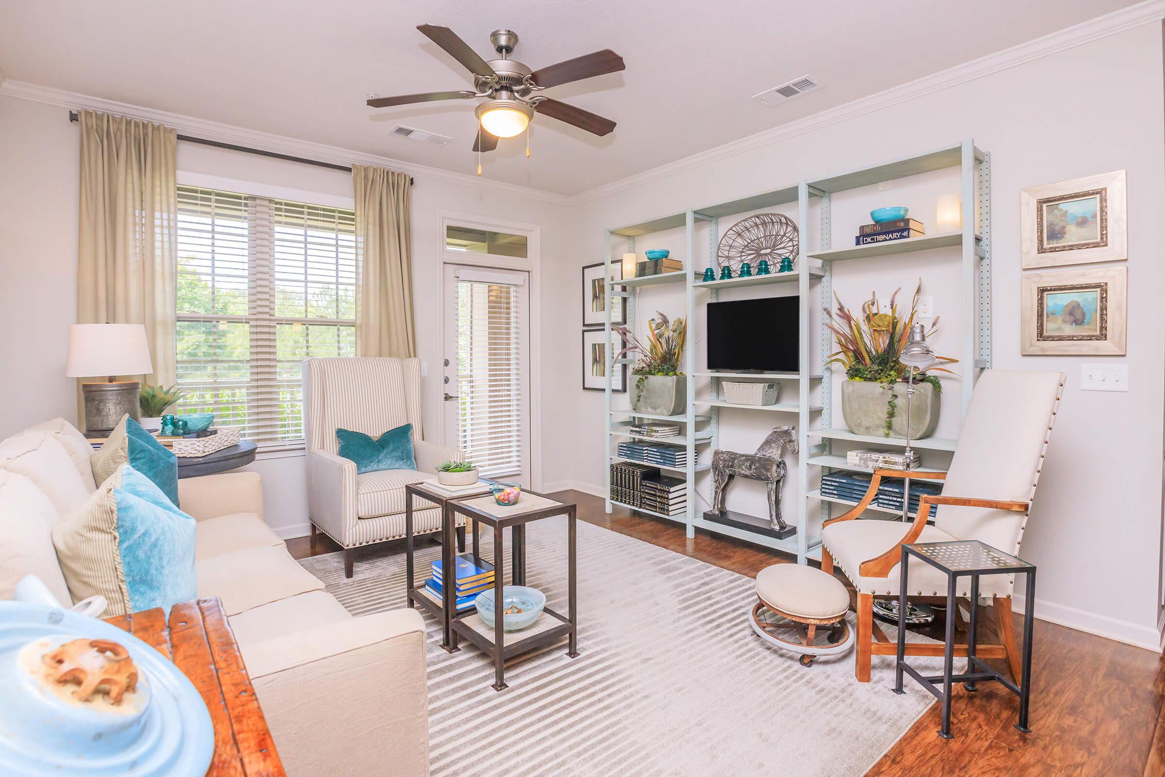 A bright and inviting living room featuring a light-colored sofa with blue accents, a small coffee table, and a stylish armchair. A bookshelf with decorative items and a TV occupies one wall, while large windows provide natural light. The space has a cozy, modern feel with hardwood flooring and soft curtains.