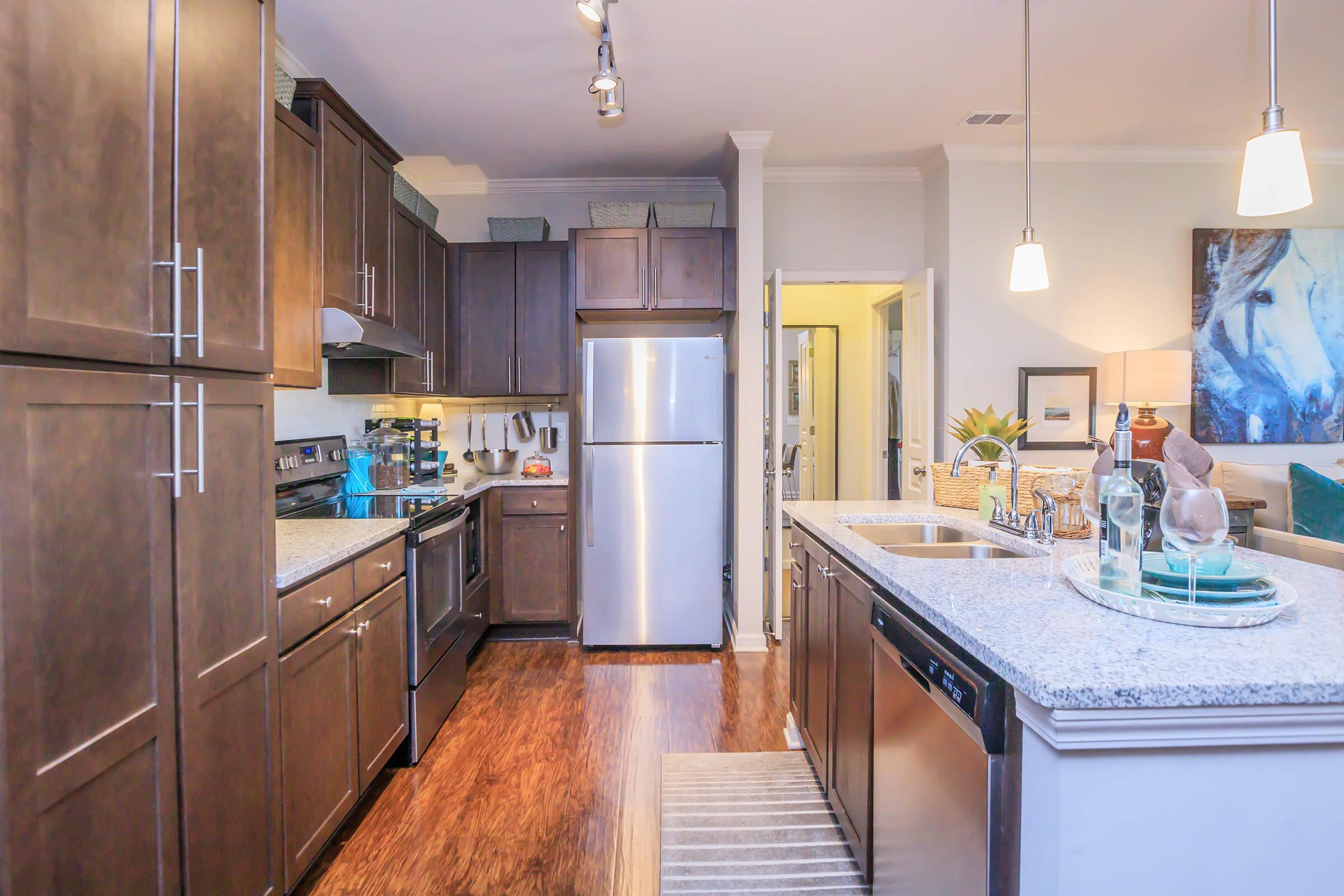A modern kitchen featuring dark wood cabinets, stainless steel appliances, and a granite countertop. The area includes a sink, oven, and refrigerator, with a cozy dining area visible in the background. Soft lighting and decorative elements create a welcoming ambiance.