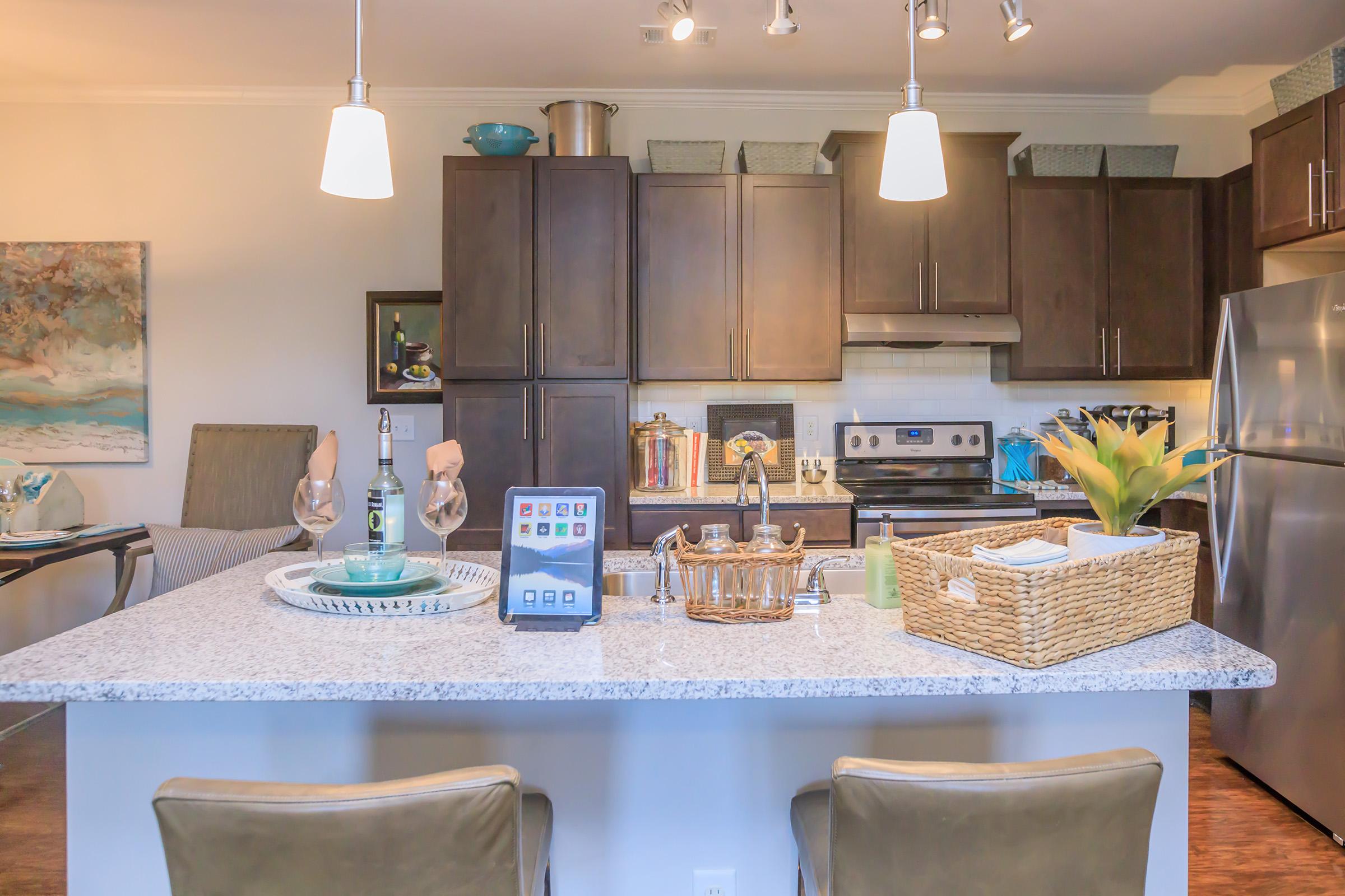 Modern kitchen featuring dark wood cabinets, stainless steel appliances, and a granite countertop. A decorative tray with a tablet, glasses, and a plant sits on the island. Pendant lights illuminate the space, which includes a cozy seating area with art on the walls.
