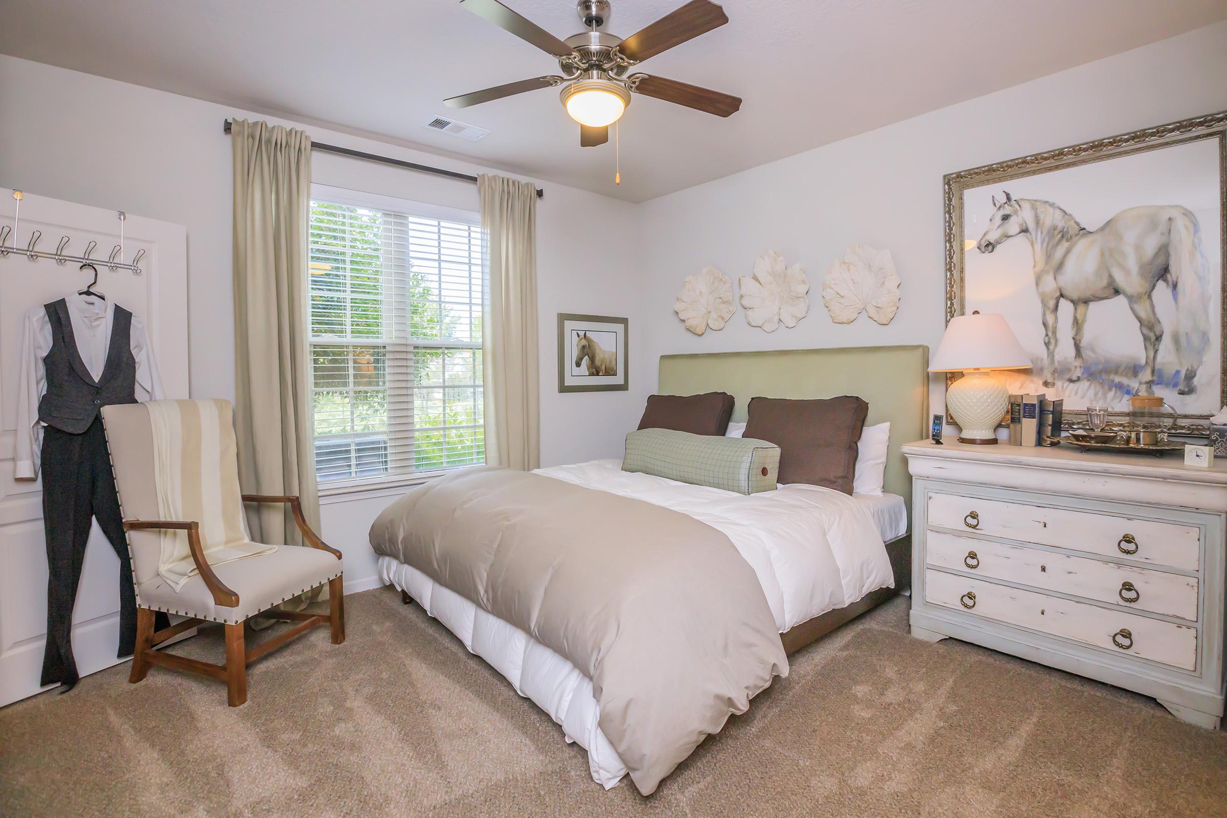 A cozy bedroom featuring a neatly made bed with beige and brown bedding, a decorative horse painting on the wall, a wooden dresser, a soft armchair, and a fan on the ceiling. Natural light streams through the window, adorned with light curtains, creating a warm and inviting atmosphere.