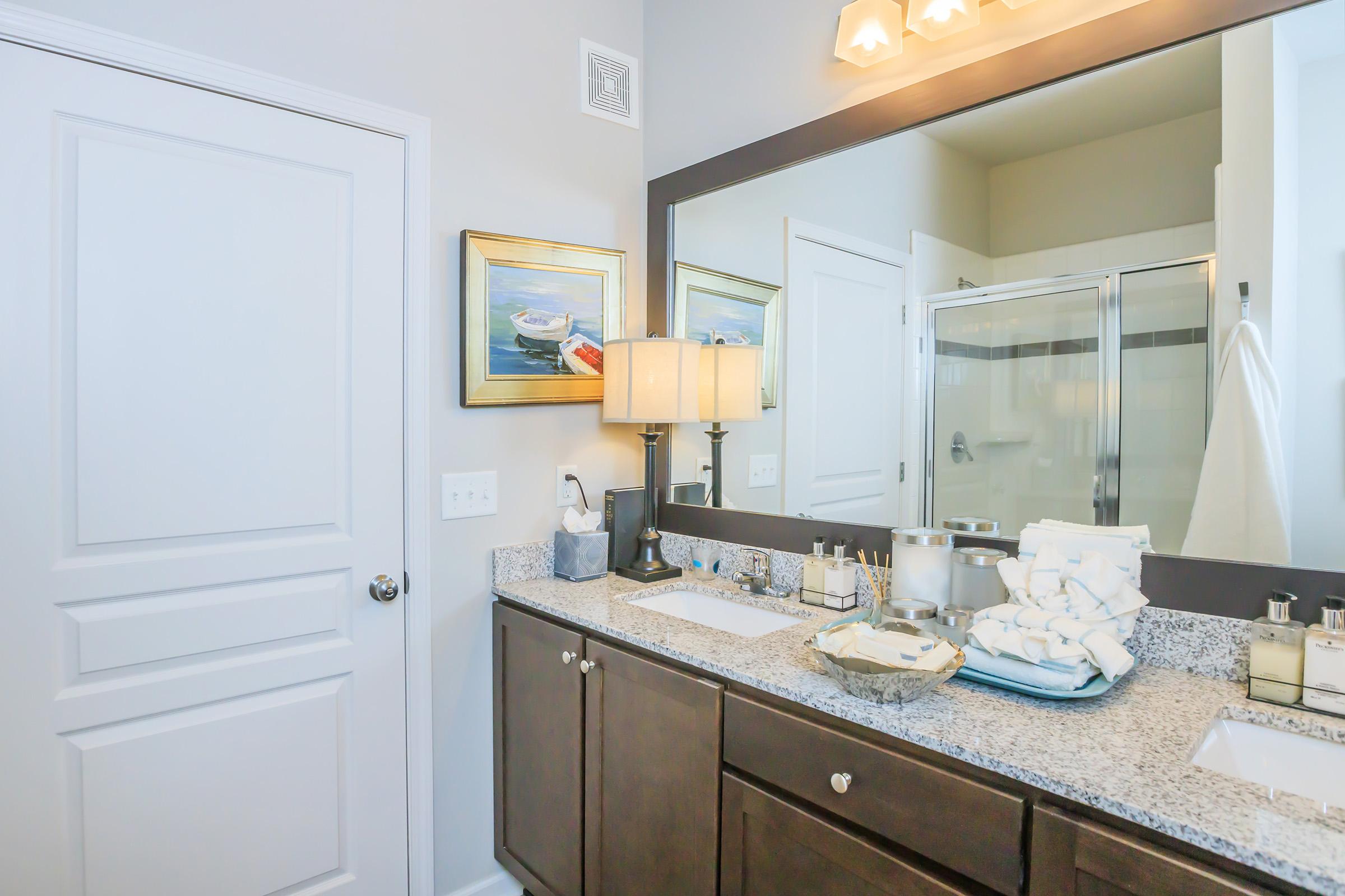 A modern bathroom featuring a large mirror above a dark wood vanity with granite countertop. There are neatly stacked white towels, a decorative lamp, and personal care items. A glass shower enclosure is visible, along with artwork of boats on the wall. The walls are painted a light color, enhancing the bright atmosphere.