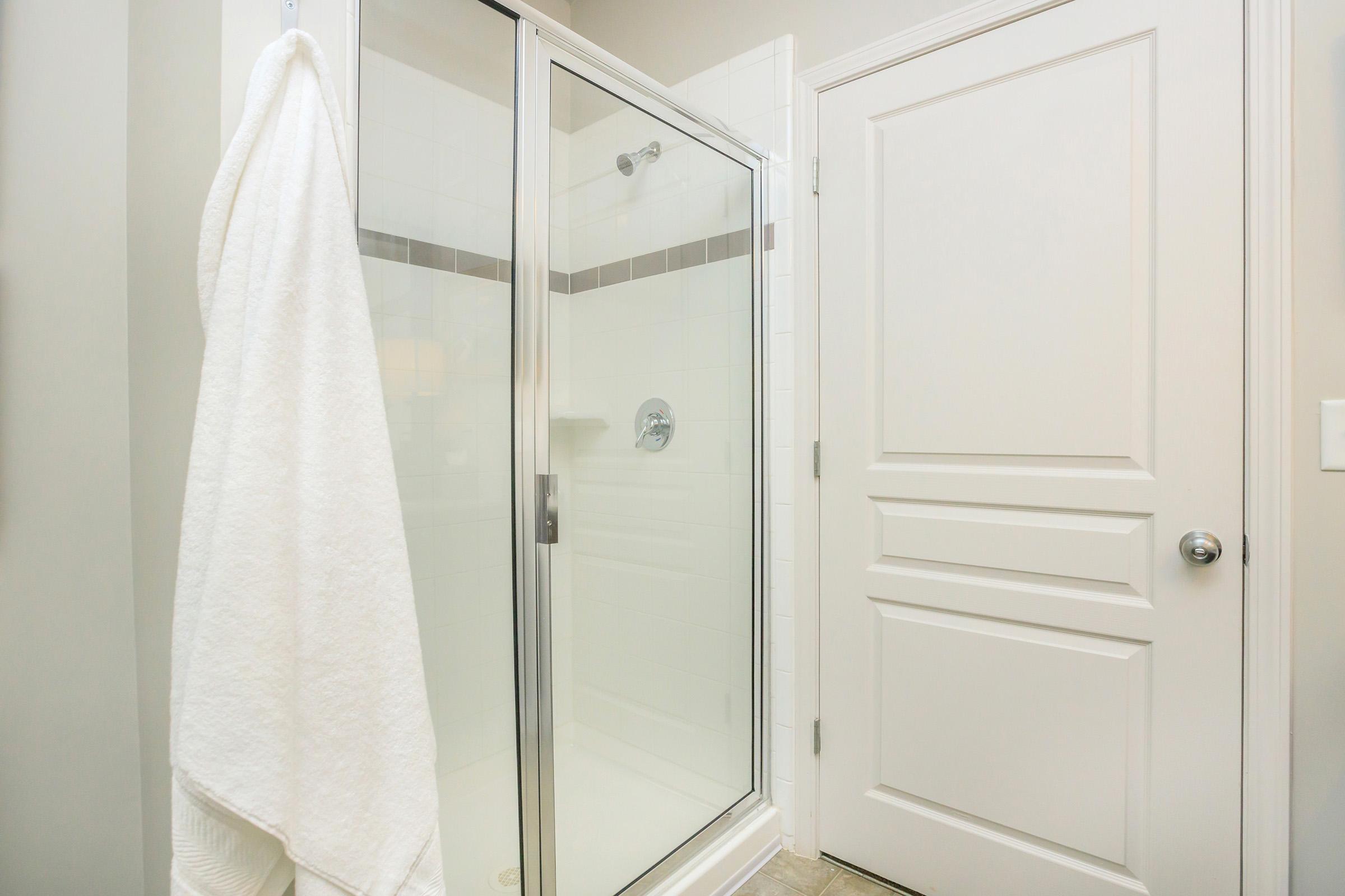 A modern bathroom featuring a glass shower enclosure with a silver handle, a white towel hanging on the door, and a closed white door with a sleek design. The walls are light-colored and the floor is tiled, creating a clean and fresh atmosphere.