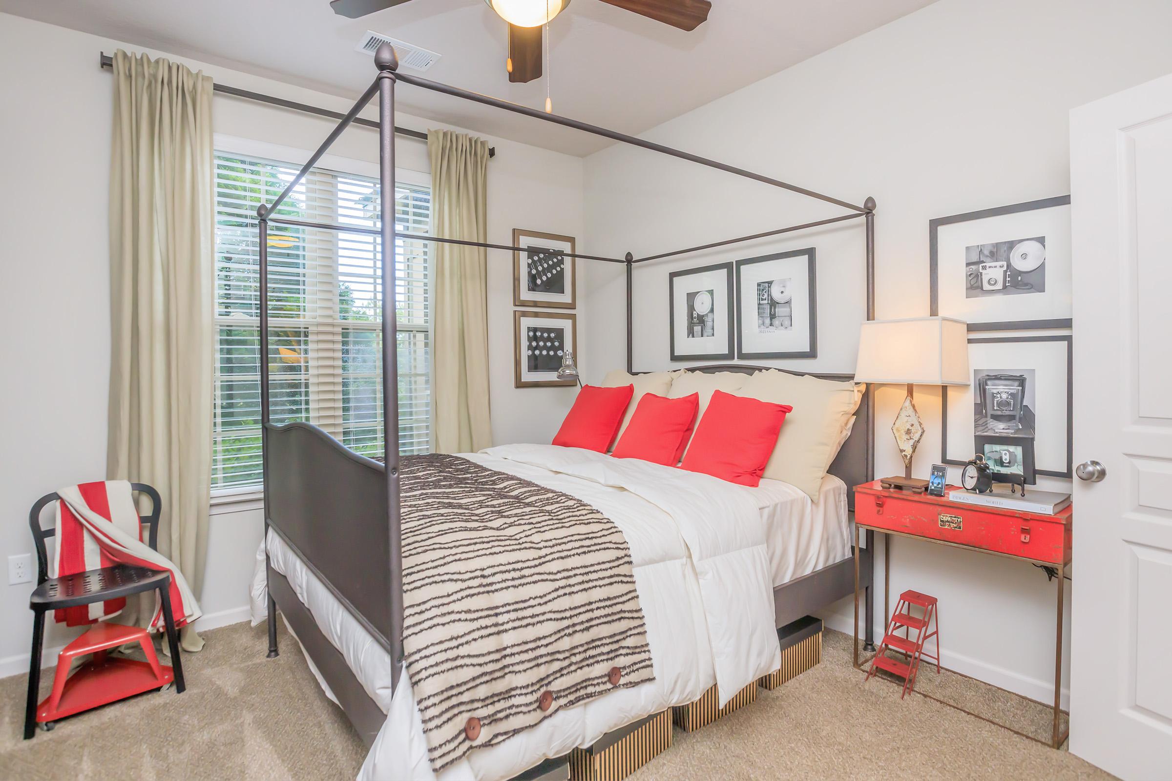 A cozy bedroom featuring a four-poster bed with red and white bedding, complemented by decorative pillows. The room has light-colored walls, a window with beige curtains, and framed artwork on the wall. A red nightstand with a lamp sits beside the bed, and there’s a black chair and a small decorative ladder in the corner.