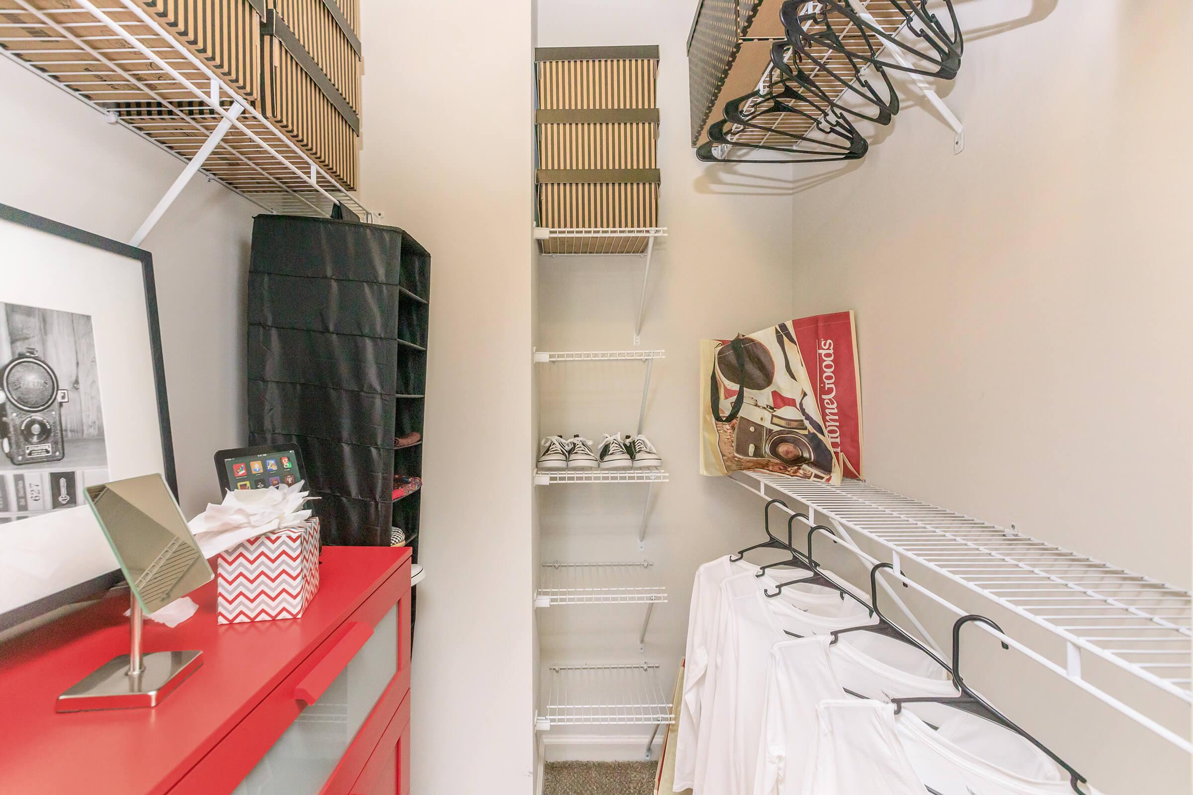 A well-organized closet featuring white clothing hung on wire shelves, a red storage unit with decorative boxes, a mirror, and a tablet on top. Additional shelves are empty, and a pair of sneakers sits on the floor. The overall space is clean and minimalistic, maximizing storage efficiency.