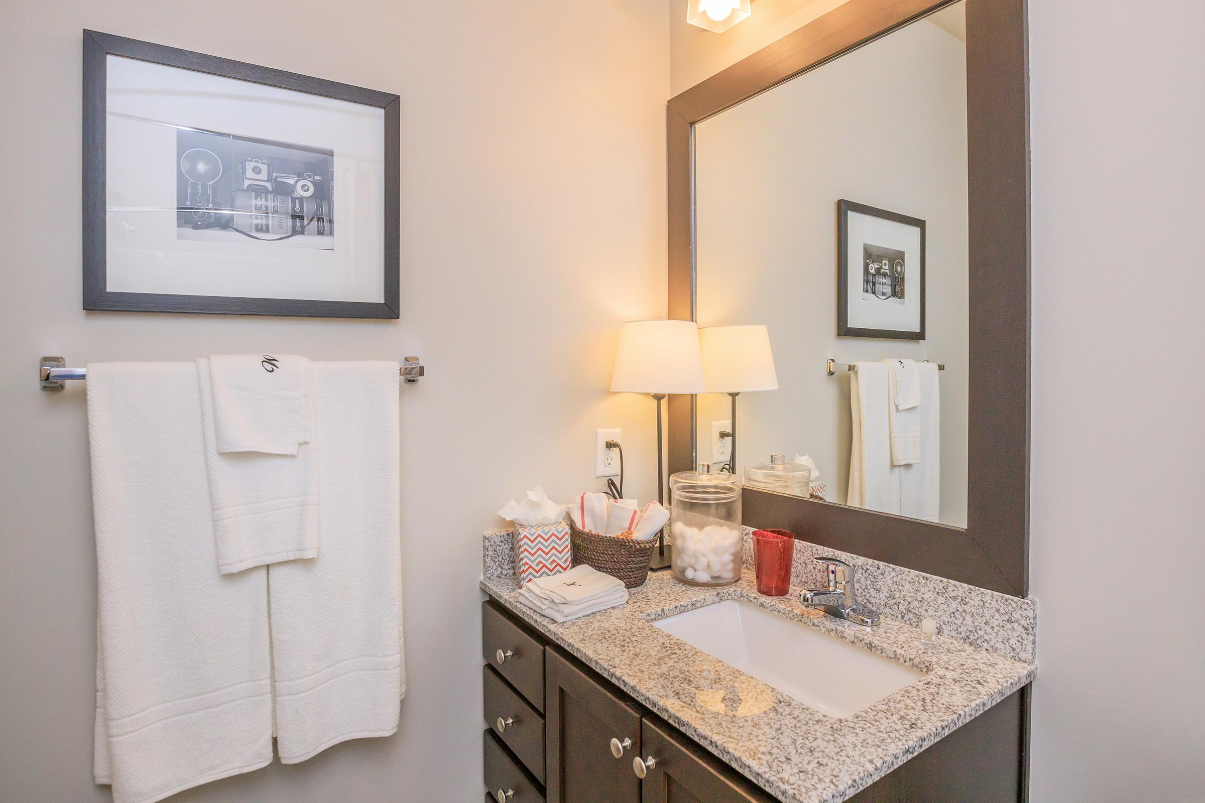 A modern bathroom featuring a stylish sink with a granite countertop, neatly arranged toiletries, a lamp, and towels hanging on a rack. A decorative mirror and framed artwork add character to the space. Cotton balls and other essentials are organized in containers, enhancing the room's functionality and aesthetic.