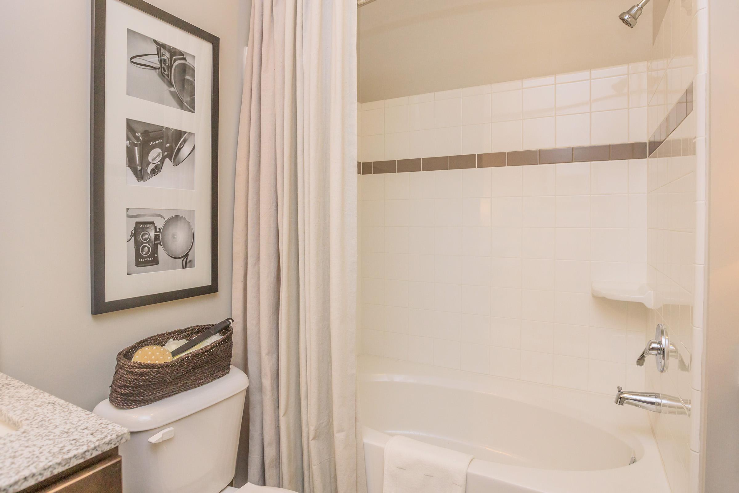 A bathroom featuring a white tiled shower and bathtub, a decorative black-framed photograph on the wall, a beige shower curtain, a granite countertop with a woven basket on top of a white toilet, and a white towel hanging nearby. The space is well-lit and clean.