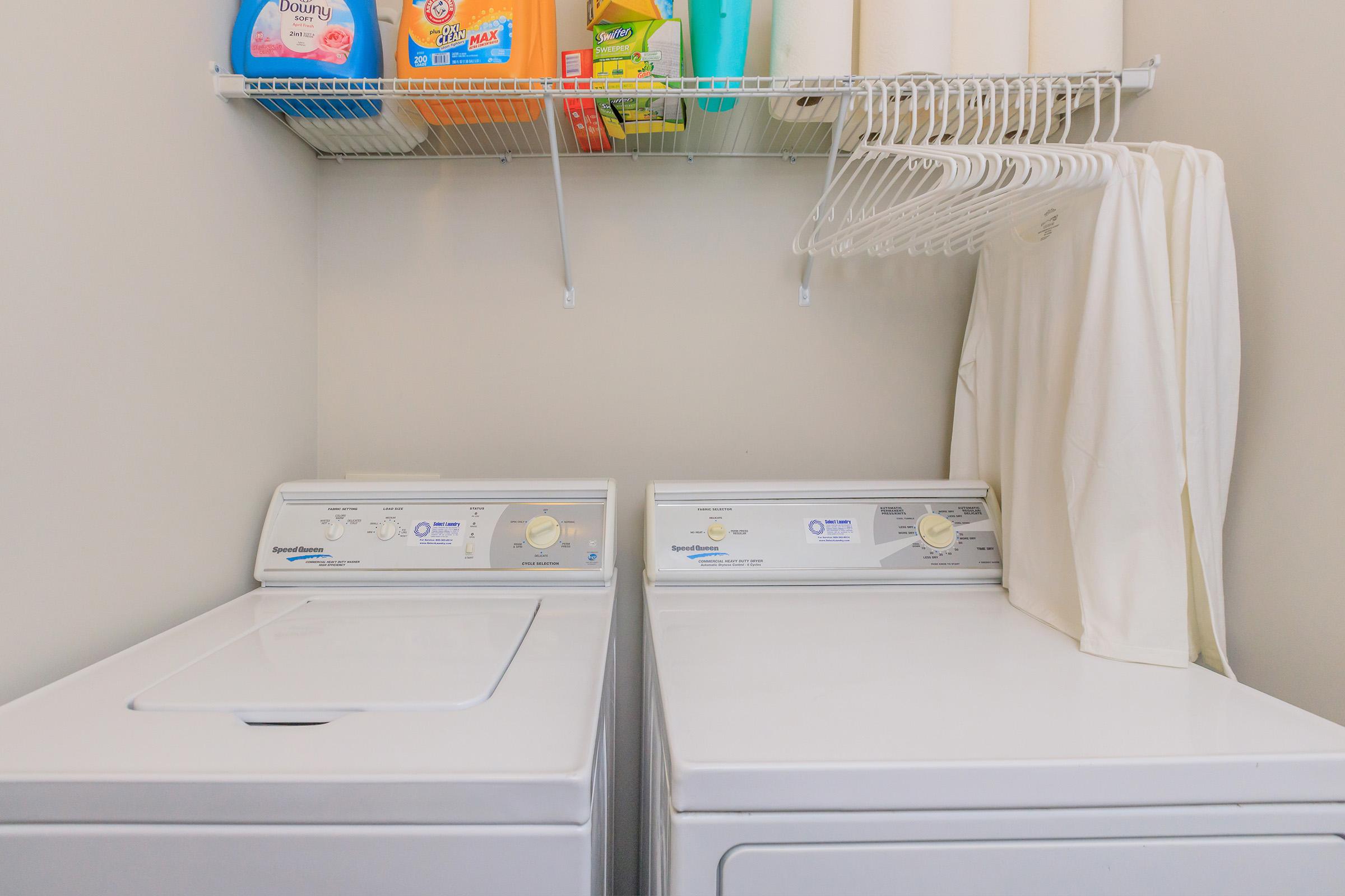 A laundry room featuring a washing machine and dryer side by side. Above them, a shelf holds laundry detergent and fabric softener. White hangers are neatly arranged on a rack, and a white garment hangs nearby, adding to the organized and clean appearance of the space.