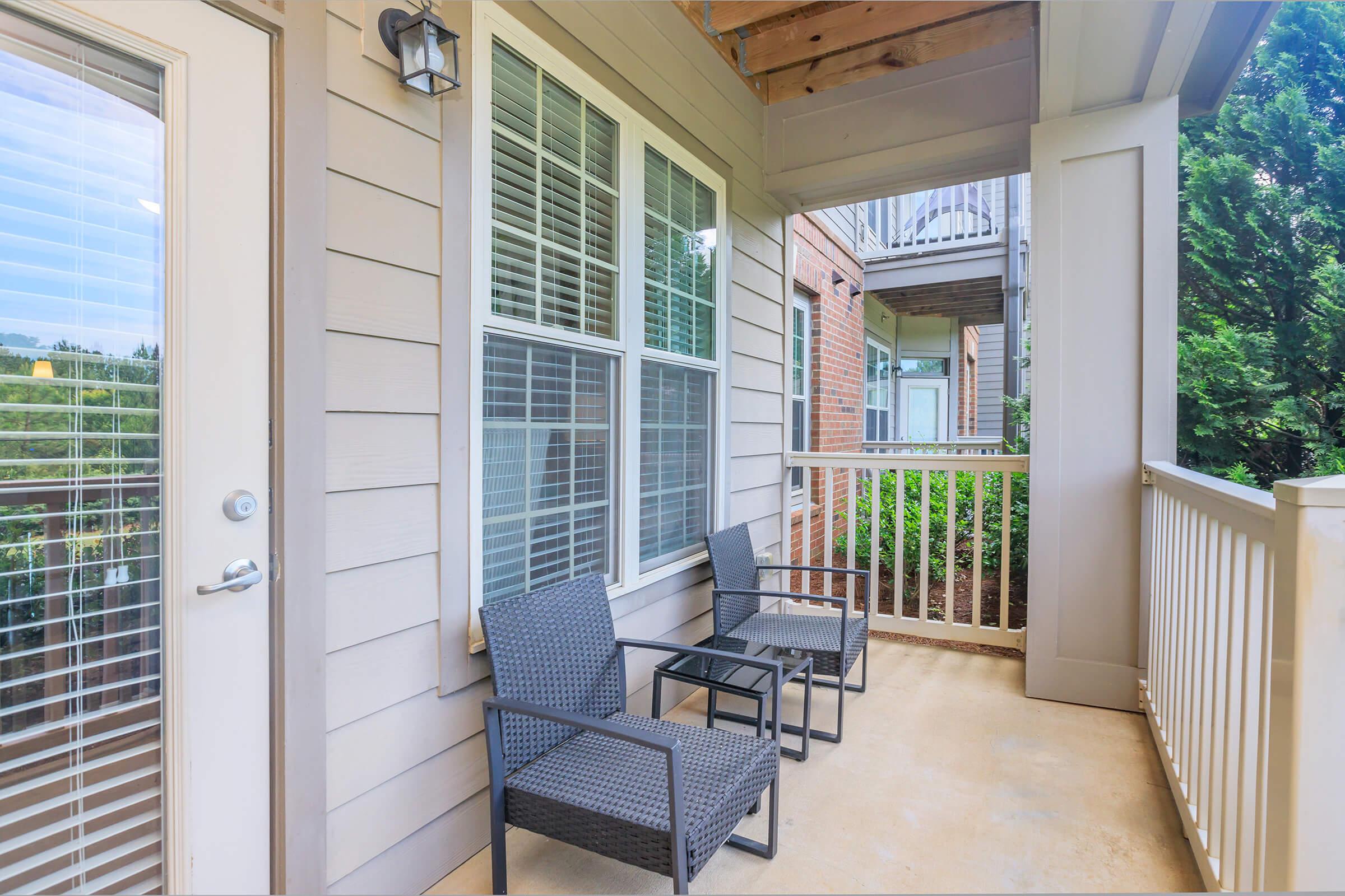 A balcony with two wicker chairs and a small table, surrounded by greenery. The building features light-colored siding and large windows with blinds. The patio is covered, creating a cozy outdoor space.