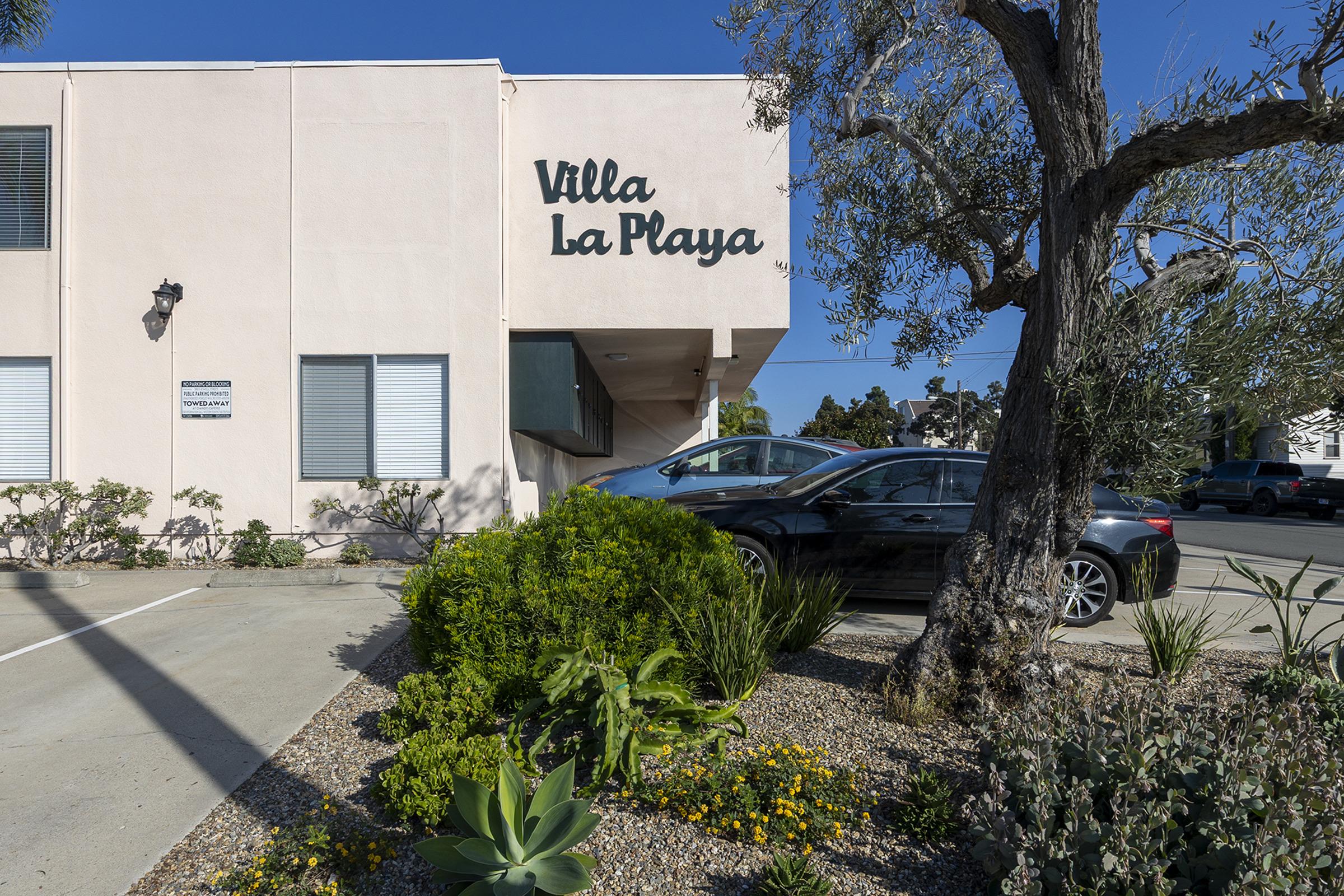 Exterior view of a building with the sign "Villa La Playa." The building features a light pink facade, parked cars in front, and landscaped greenery including various plants and shrubs. A tree is visible to the right, adding to the scenic atmosphere on a clear, sunny day.