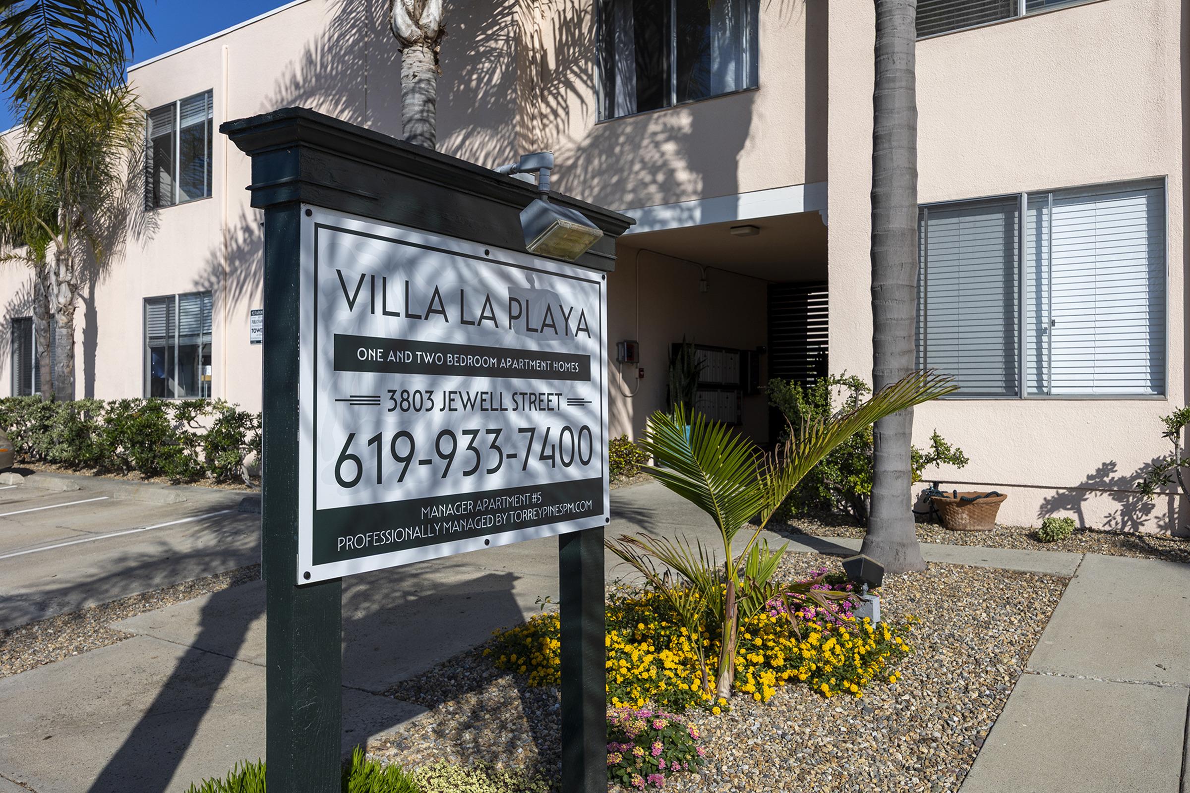 Sign for Villa La Playa, listing one and two-bedroom apartments for rent. Address is 3805 Jewell Street, with a contact number of 619-933-7400. Surrounding the sign are palm trees and colorful flowers, with the building visible in the background.