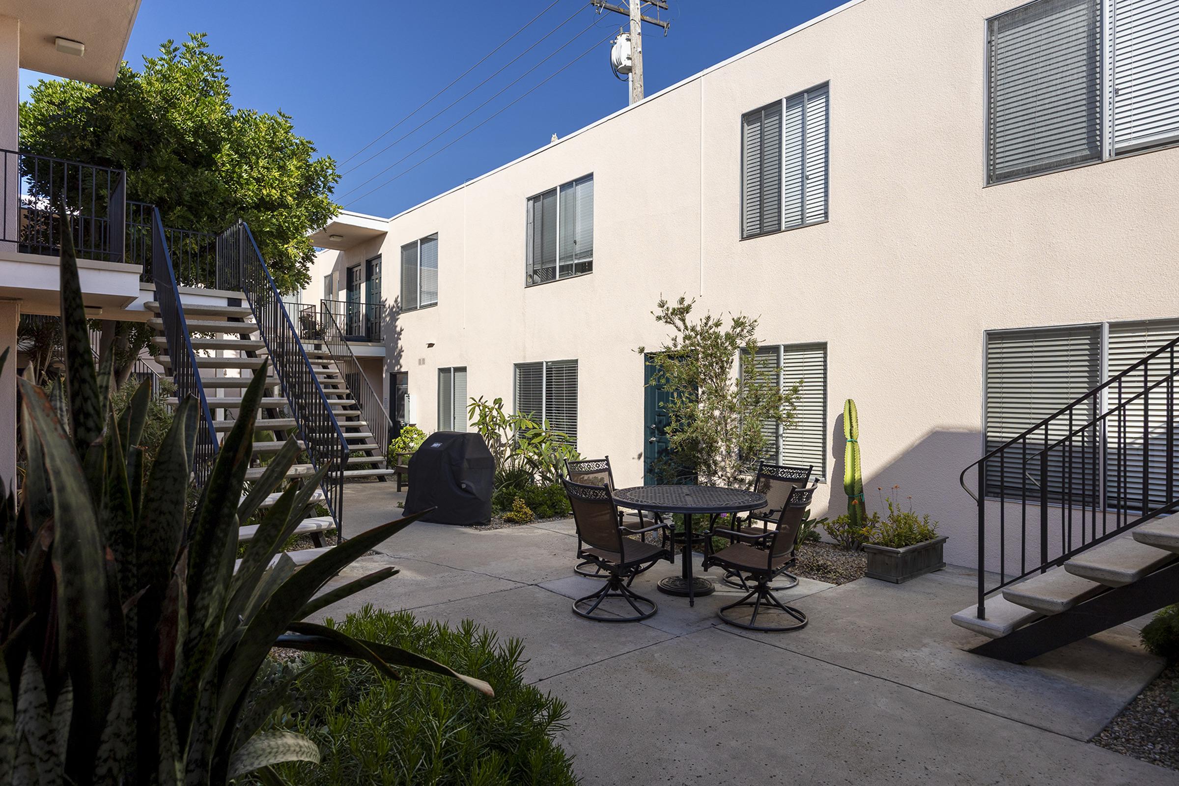 A view of a courtyard featuring two-story apartment buildings with a palm tree and cactus. A round table and chairs sit on a concrete patio surrounded by greenery. Stairs lead to the upper level of the buildings, under clear blue skies.