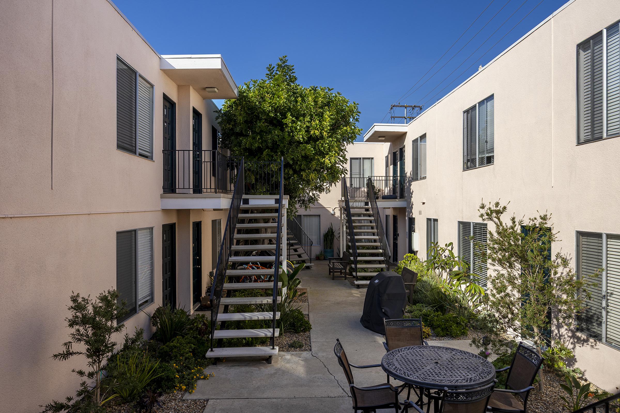 A view of a modern apartment complex courtyard featuring two buildings with balconies, surrounded by greenery. The courtyard has a staircase leading to upper levels, outdoor furniture, and a grill. The sky is clear and blue, adding natural light to the scene.
