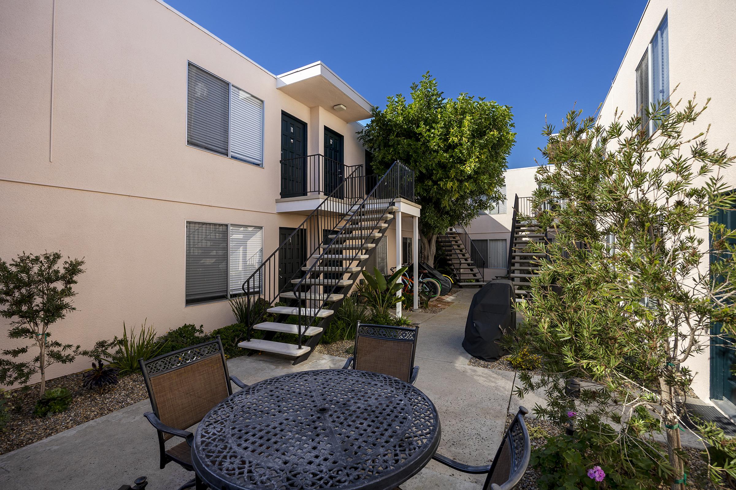 A courtyard scene featuring two buildings with light-colored exteriors. There are staircases leading to upper levels on both sides. A round metal table and chairs are positioned in the center, surrounded by shrubs and small plants. The sky is clear and blue, creating a bright, inviting atmosphere.