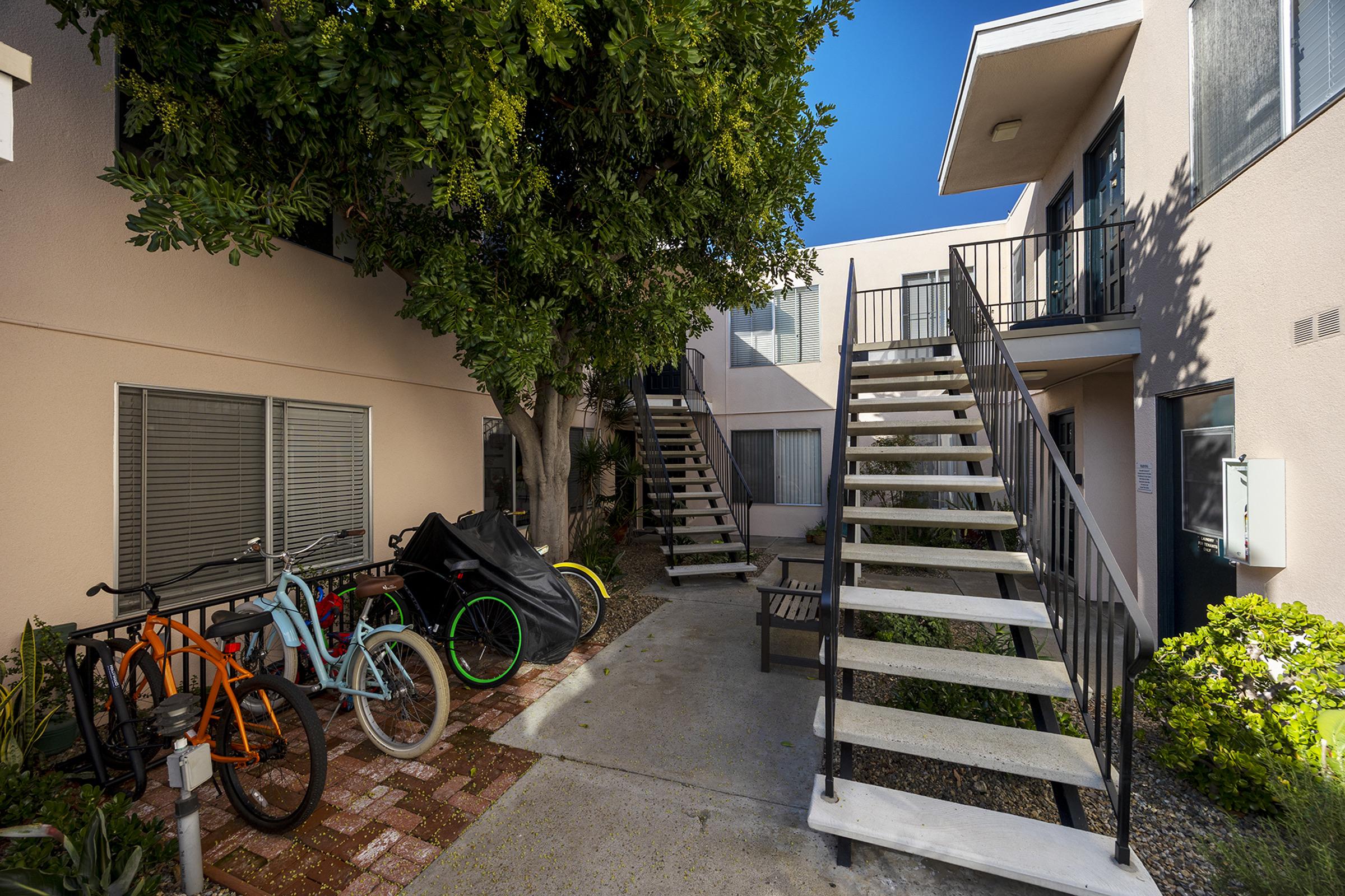 A well-lit outdoor area between two buildings featuring a stairway, a bench, and a collection of bicycles in various colors parked nearby. Lush greenery and bright blue sky add to the inviting atmosphere of the space.