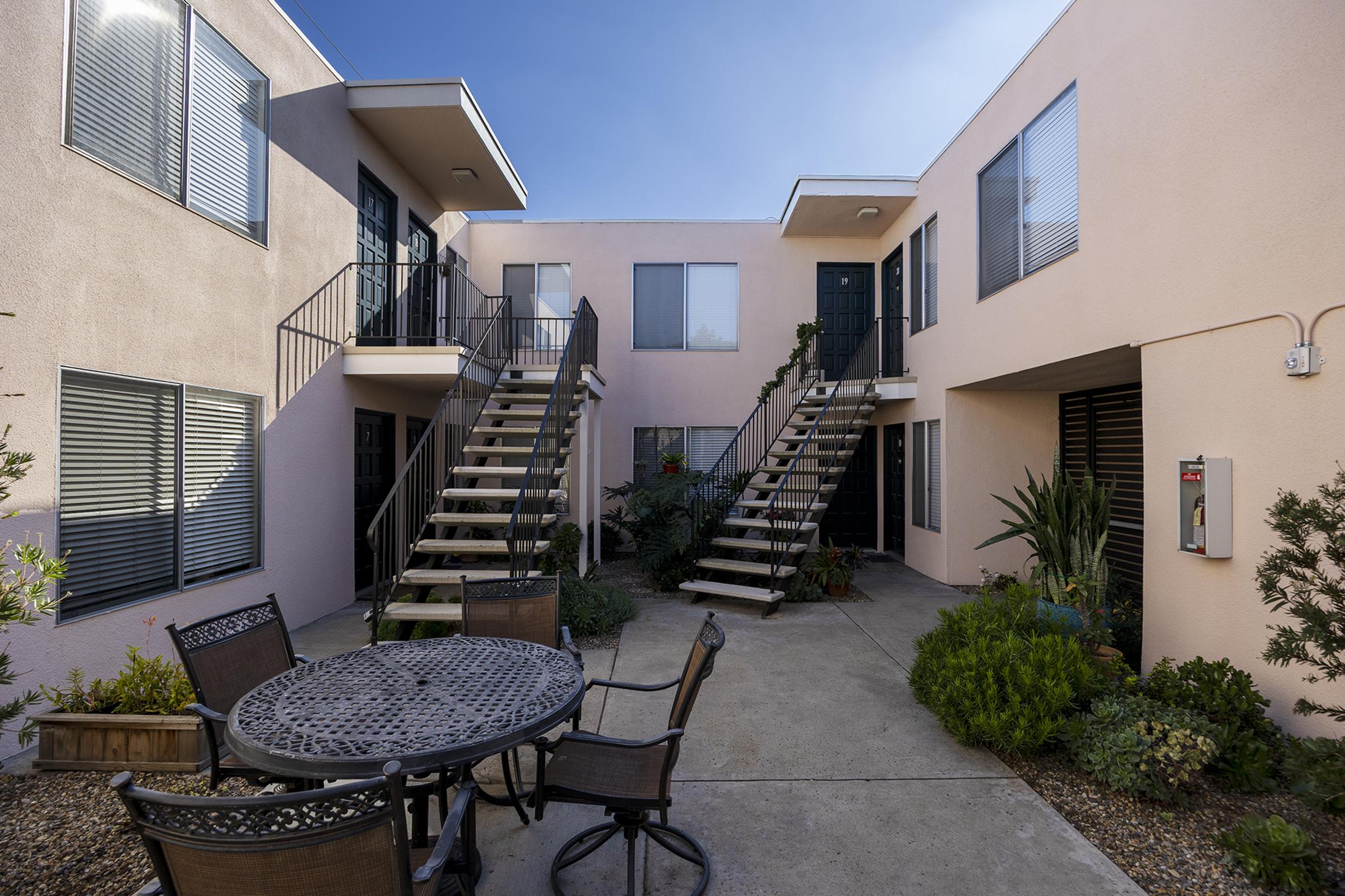 A courtyard of a multi-story apartment complex featuring two sets of stairs leading to upper units. There is a round table with chairs in the foreground, surrounded by various plants, and a clear blue sky above. The buildings have a light pink exterior and large windows.