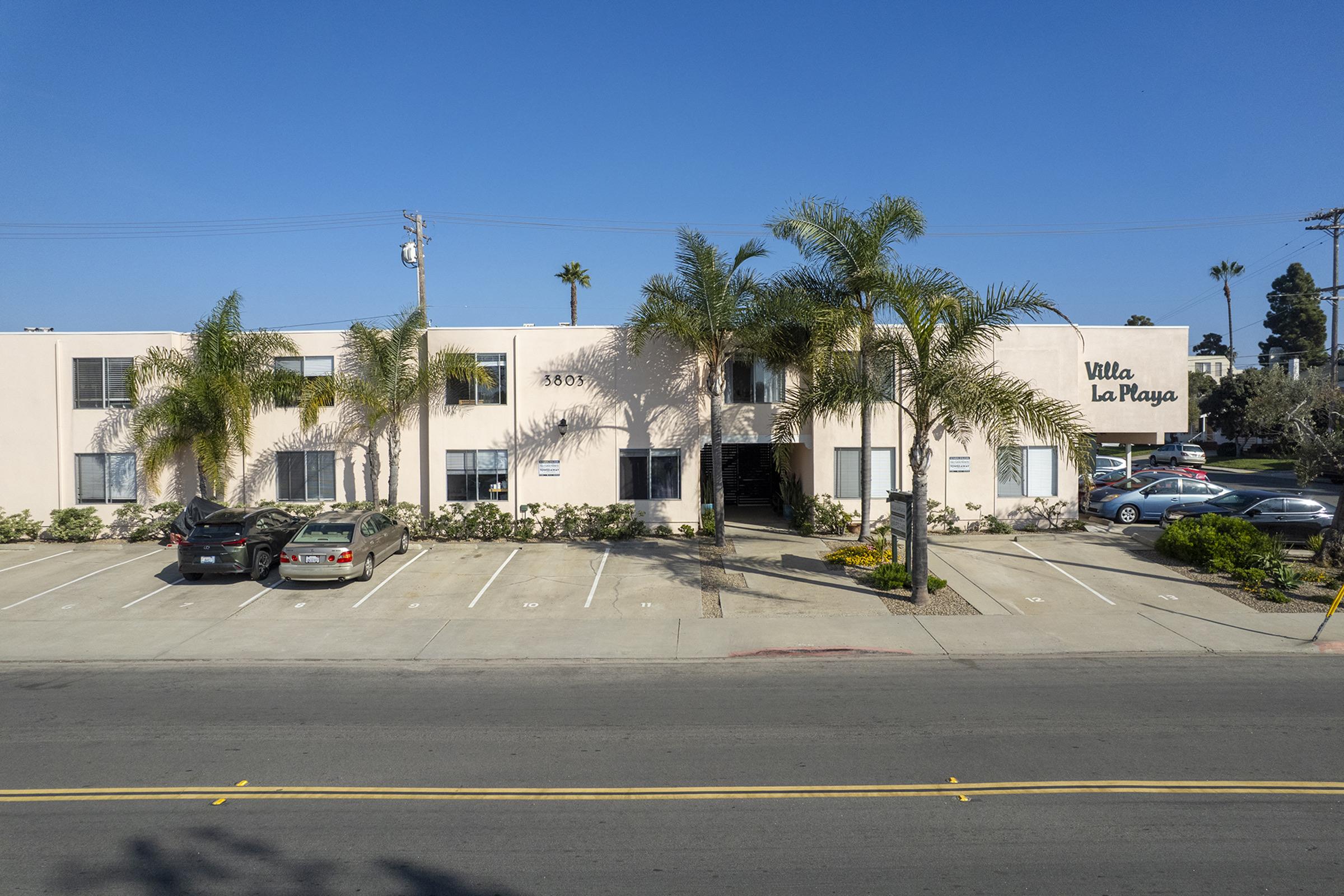 A low-rise apartment building named "Villa La Playa" with palm trees in front, situated on a sunny day. The building features multiple windows, a light exterior, and a parking area with several cars. The street in front is clear, with a blue sky and a few palm trees visible.