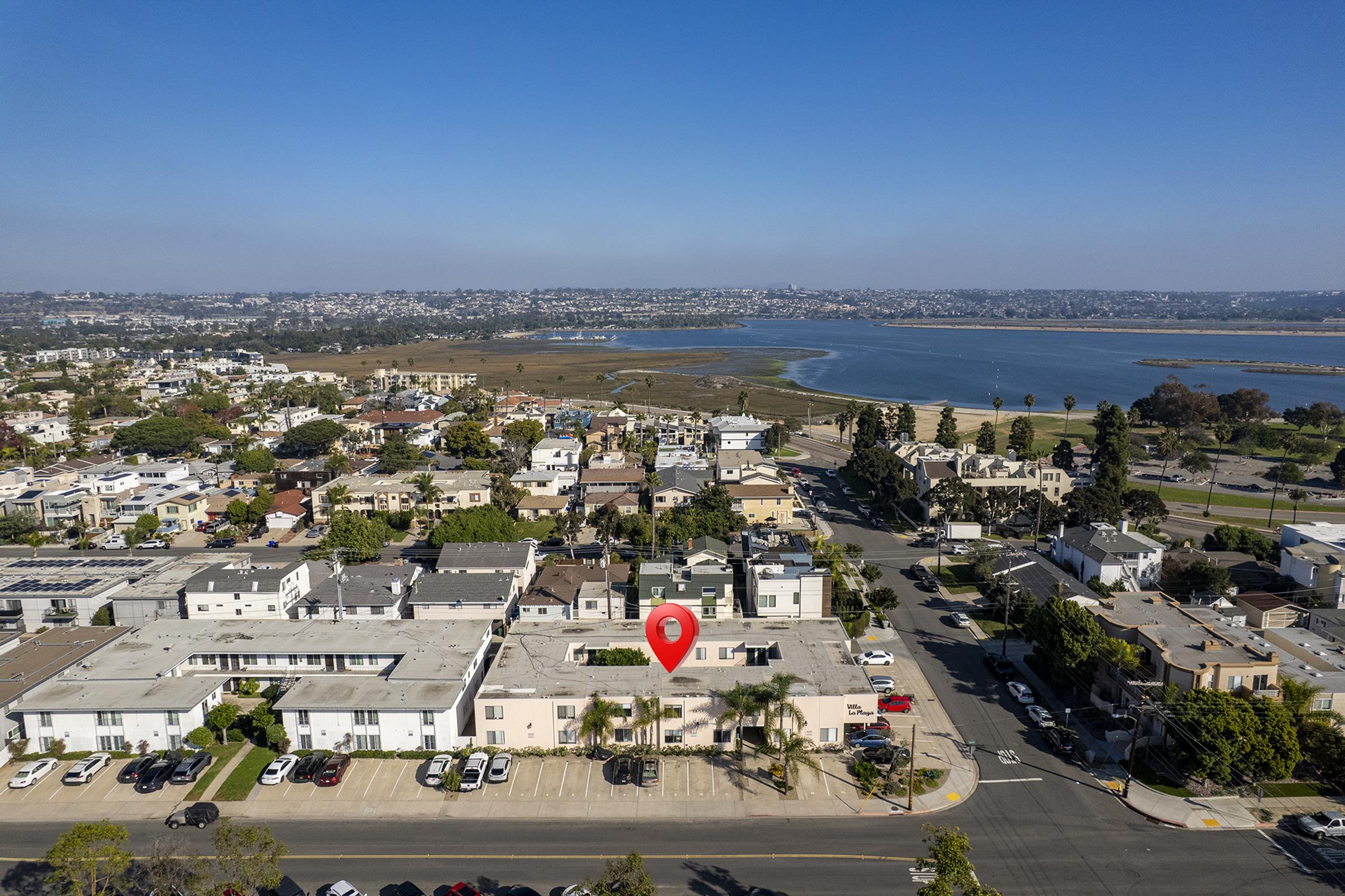 Aerial view of a coastal neighborhood featuring residential buildings, a parking lot, and a body of water in the background. A red location pin marks a specific point on a flat rooftop, with clear skies and distant cityscape visible.