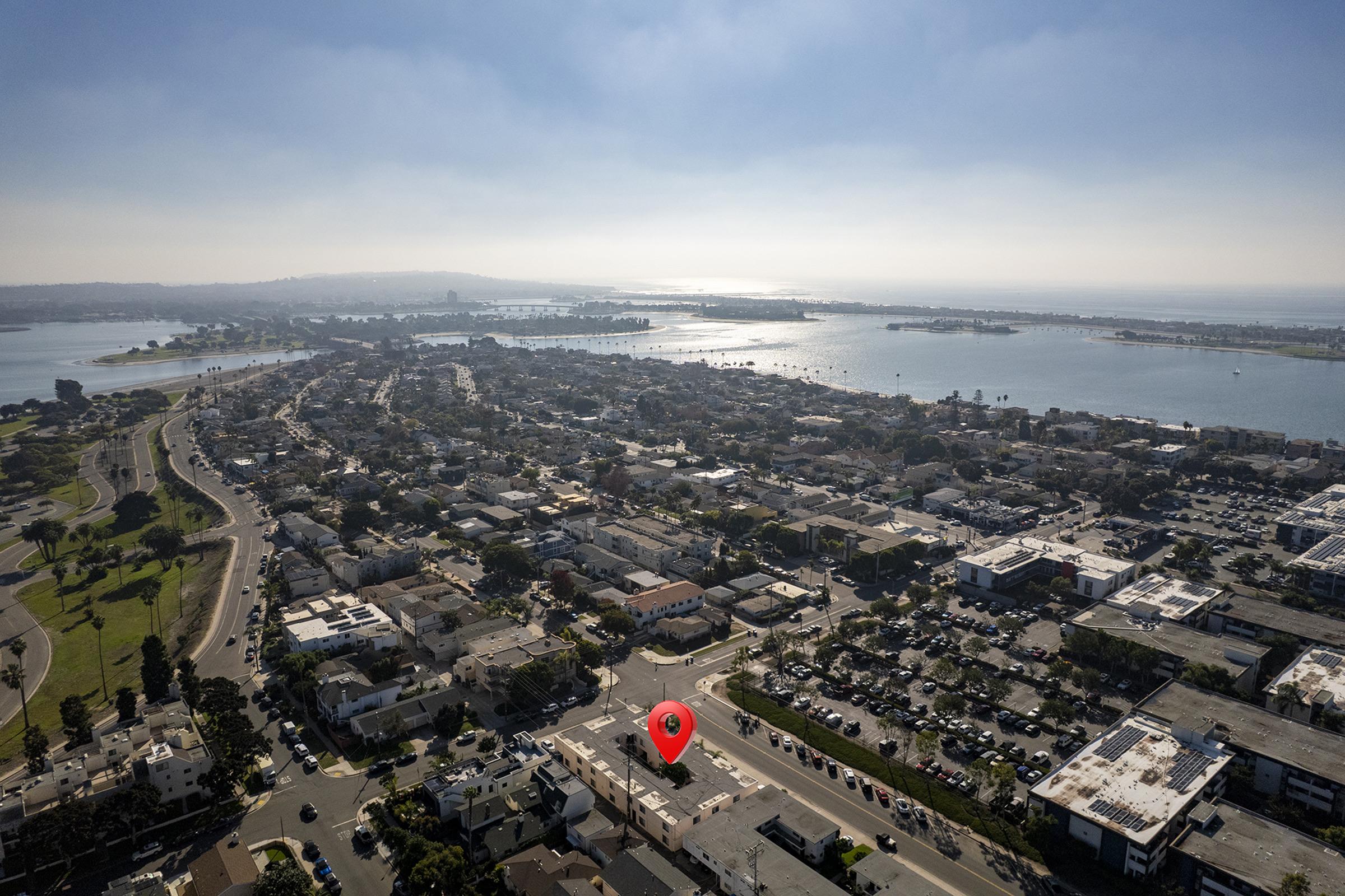 Aerial view of a coastal area featuring a residential neighborhood, roads, and waterways. A red location marker indicates a specific point of interest, with the scenic waterfront and distant hills visible in the background. The scene captures a blend of urban development and natural beauty.