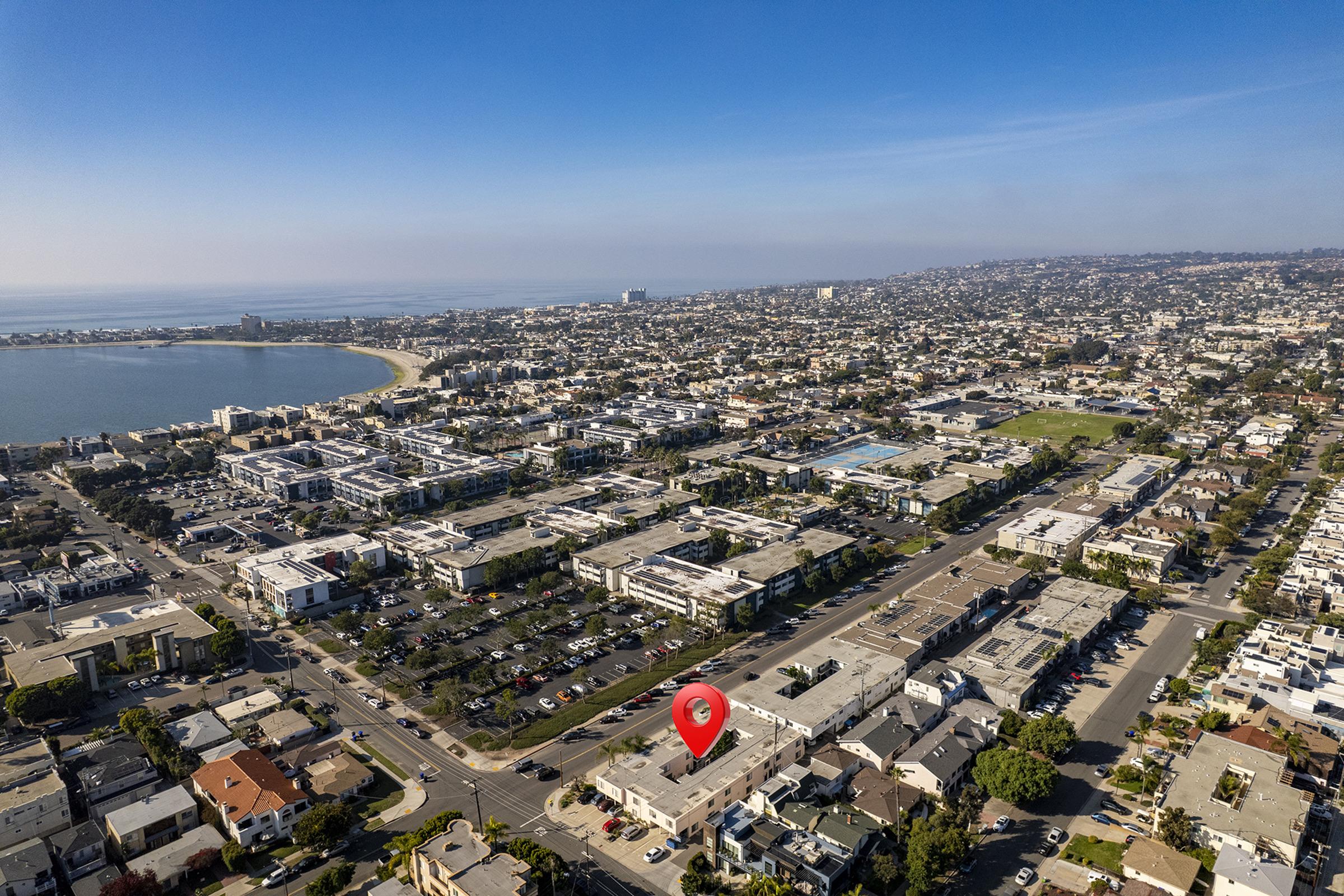 Aerial view of a coastal city showcasing residential neighborhoods, a beach coastline, and a clear blue sky. A red marker indicates a specific location among various buildings and roads, with green spaces and parking visible in the area. The ocean is in the background.