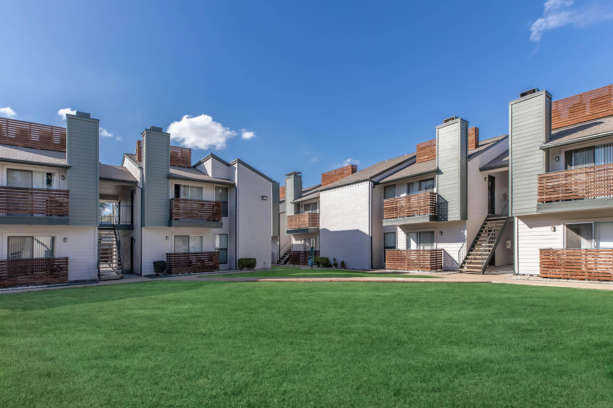 A well-maintained apartment complex featuring several buildings with balconies, surrounded by a green lawn under a clear blue sky. Some units have wooden railings, and there are stairs leading to upper-level apartments. The scene conveys a welcoming, residential atmosphere.
