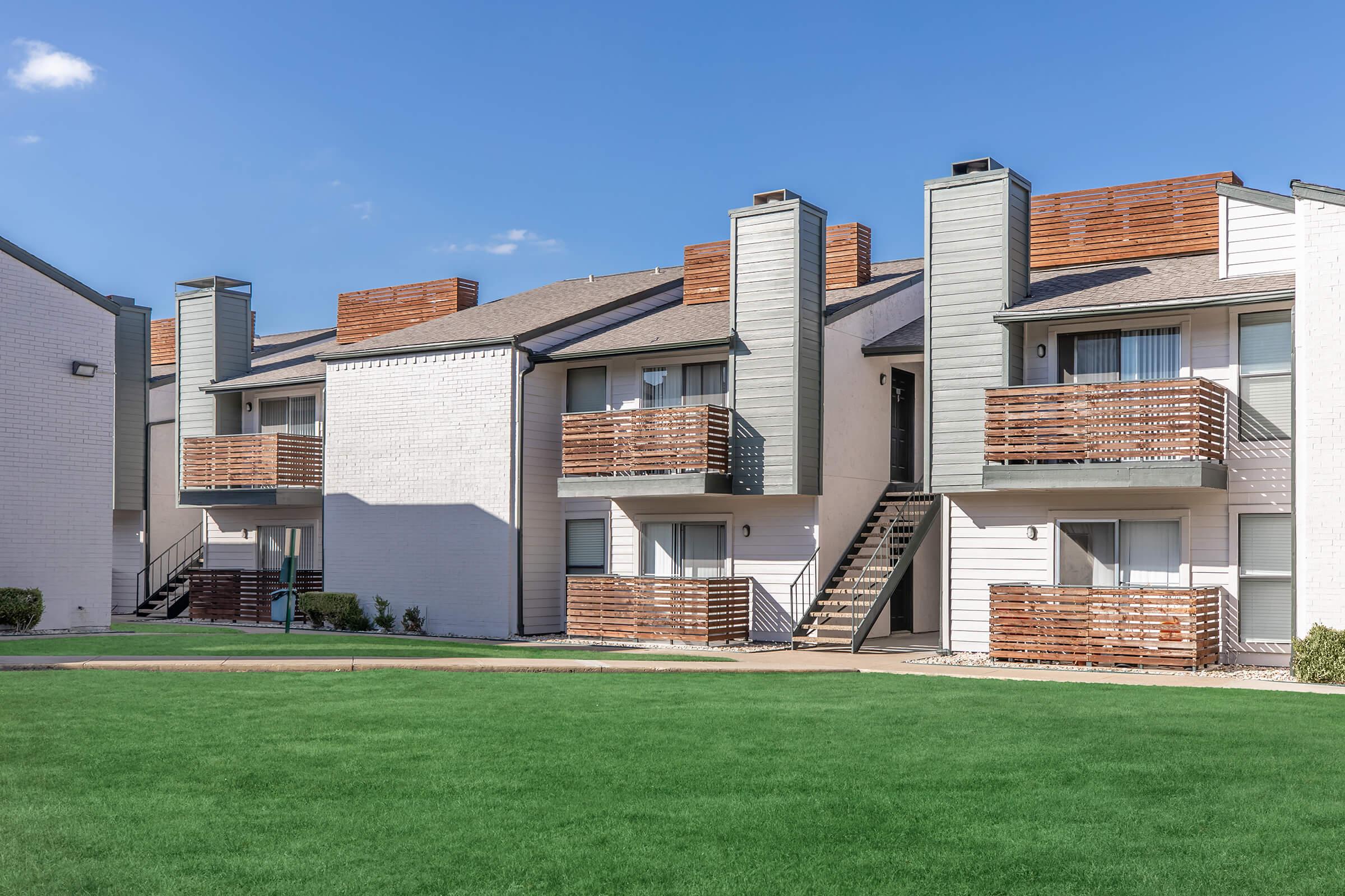 A row of multi-unit apartment buildings with two stories, featuring balconies with wooden railings. The buildings have a mix of white and gray exteriors, with some decorative wooden accents on the roofs. A well-maintained green lawn is in the foreground, and the sky is clear with a few clouds.
