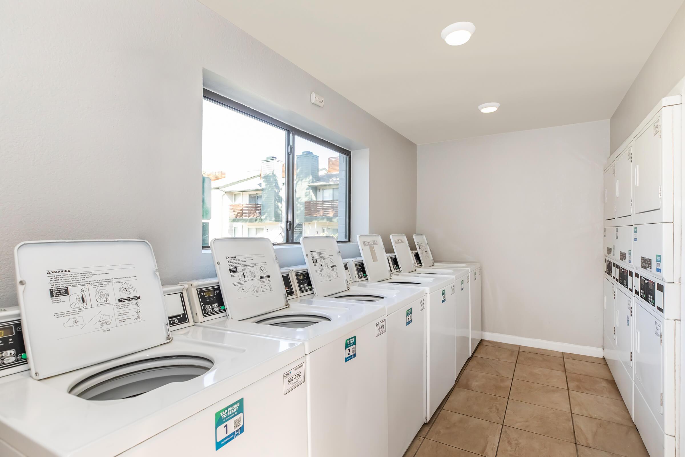 A clean and bright laundry room featuring multiple washing machines and dryers lined up against a wall, with a window allowing natural light to enter. The tiled floor and gray walls contribute to a modern, functional atmosphere suitable for laundry activities.
