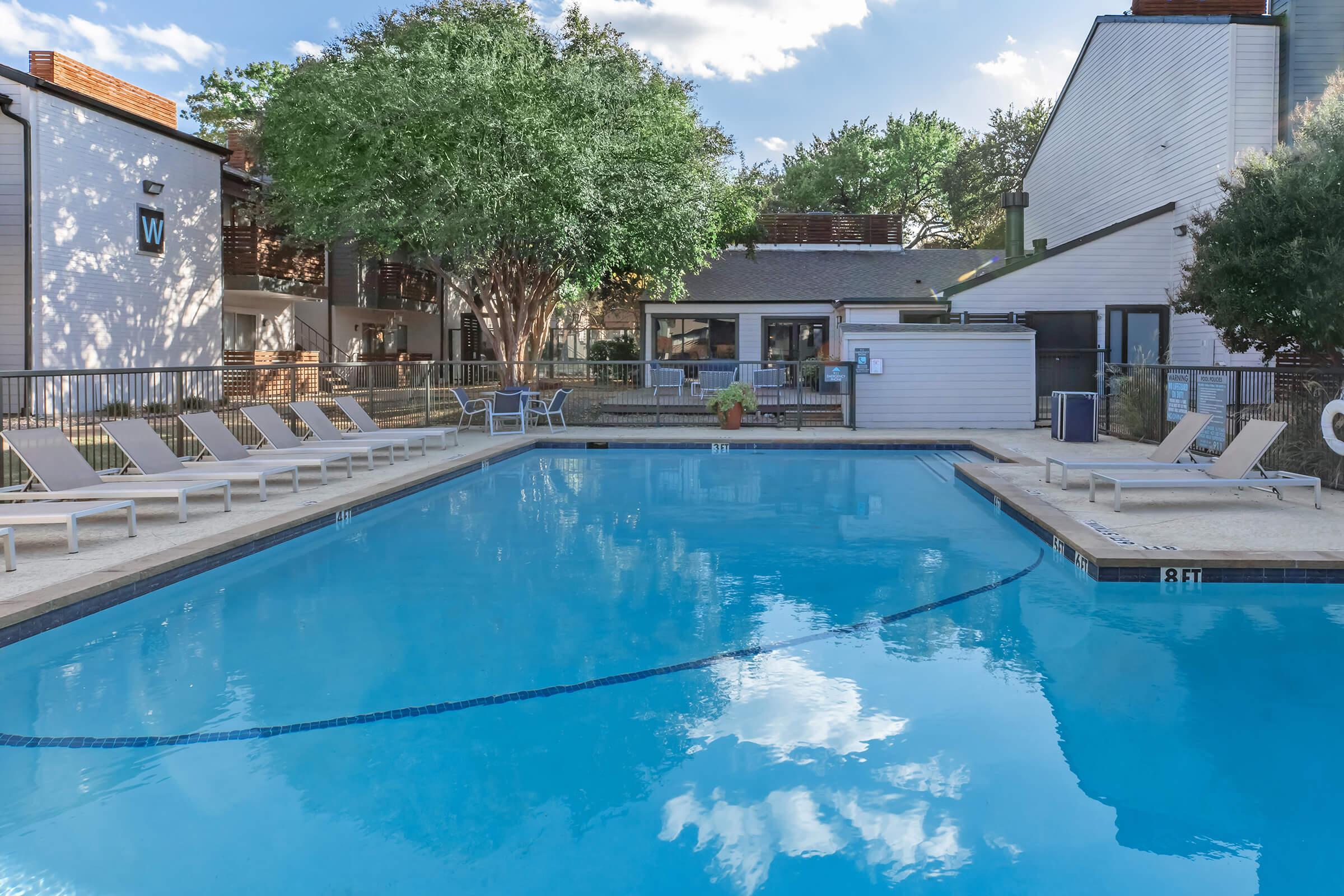 A clear blue swimming pool surrounded by lounge chairs and grassy areas. In the background, there are buildings with a mix of greenery and trees. The sky is partly cloudy, reflecting in the water. The scene conveys a relaxing outdoor space for leisure and recreation.