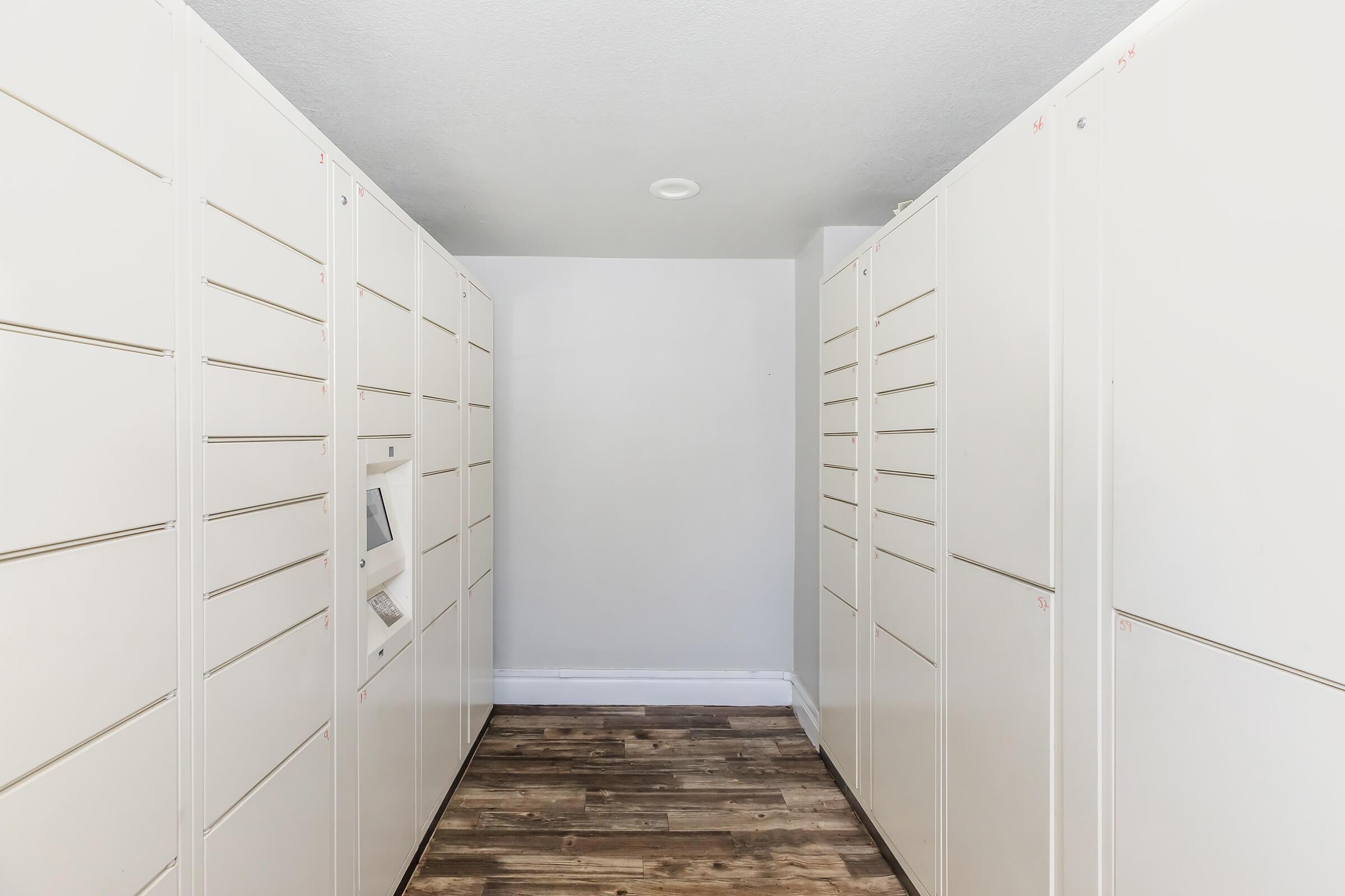 A narrow hallway with white storage lockers lining both sides. The lockers have horizontal slots for storage, and the floor is wooden with a light finish. The walls are painted a light color, and there is a panel or control unit visible on one side. The overall ambiance is clean and organized.