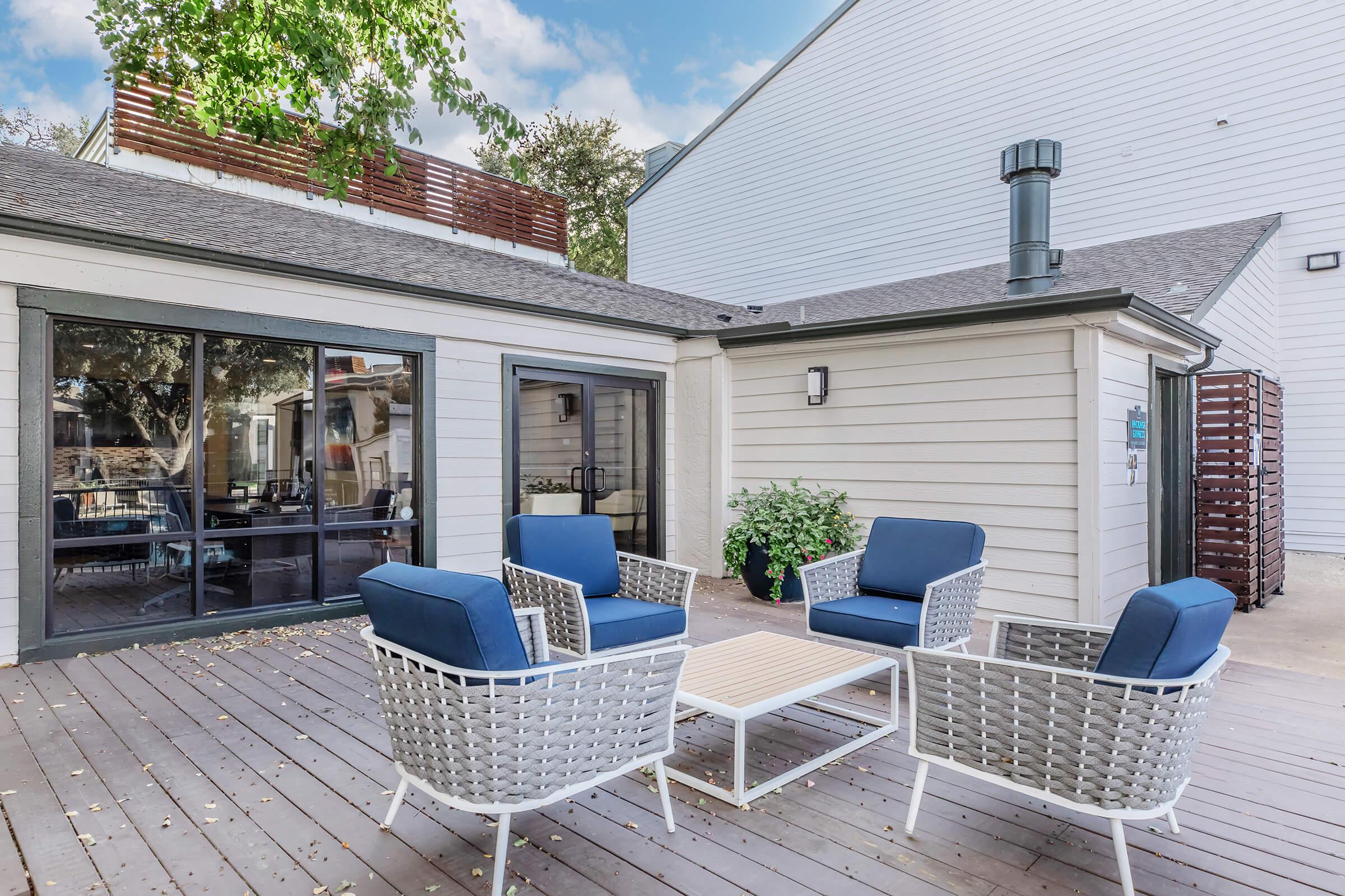 Outdoor seating area featuring four blue cushioned chairs around a wooden table on a deck. A glass door leads to an indoor space, with greenery and a chimney visible. The background includes a light-colored building and a clear sky.