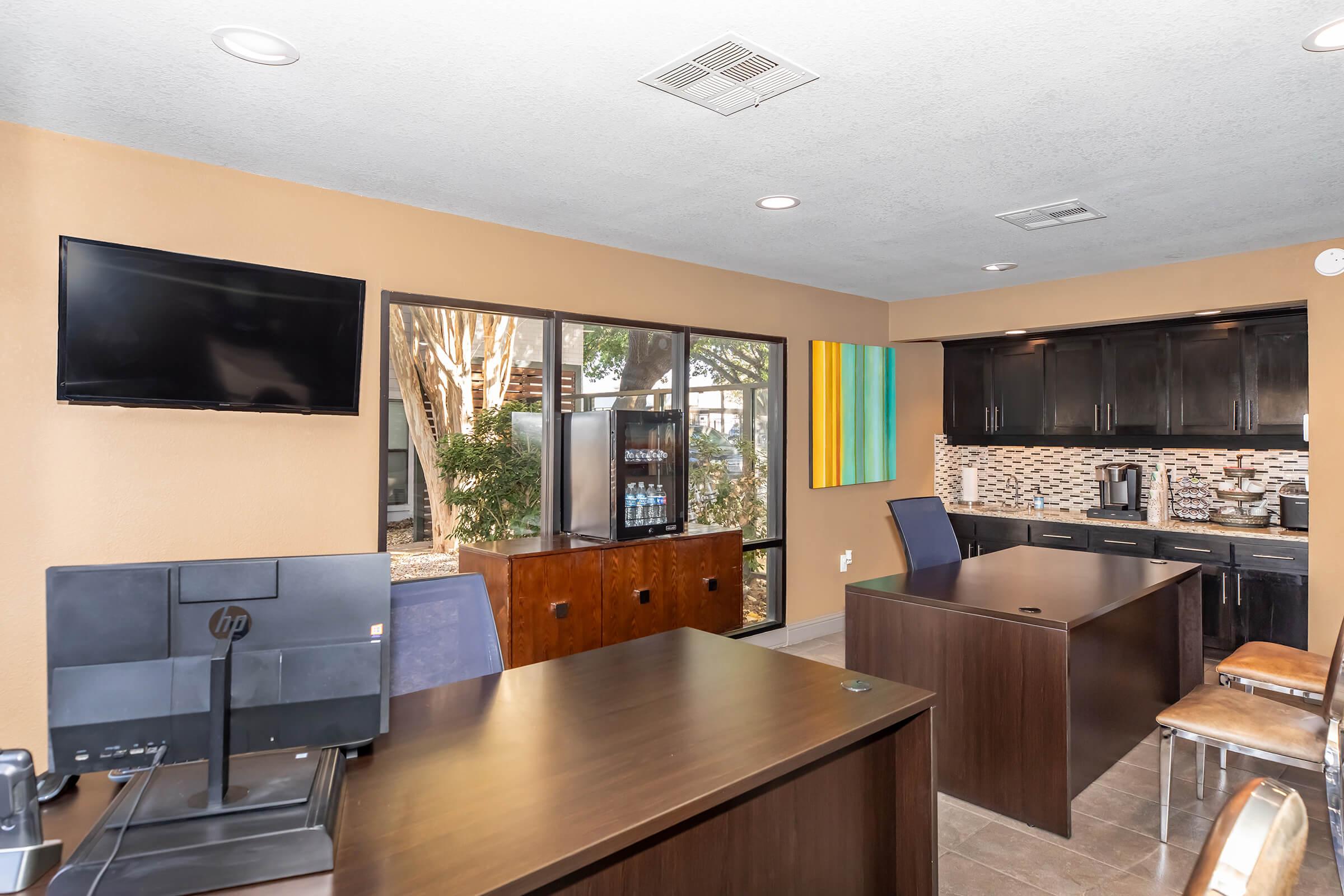 A modern office space featuring two dark wooden desks, a large window with natural light, and a flat-screen TV mounted on the wall. The decor includes an abstract painting and a small kitchenette area with a coffee maker and shelves. The atmosphere is bright and inviting, suitable for work or meetings.