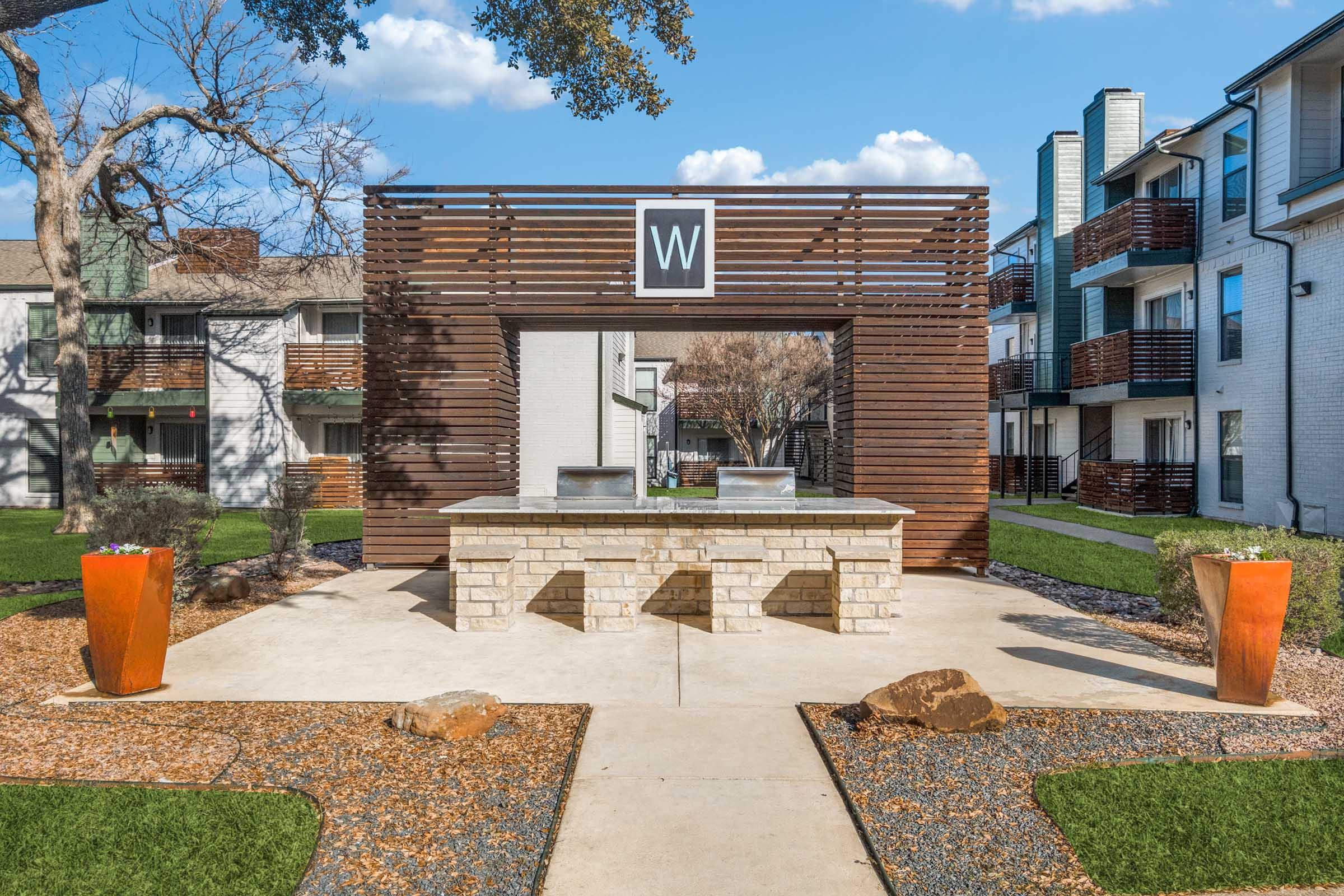 Outdoor area featuring a stone and wood structure with a large "W" logo, flanked by planters. The space includes two grills and is surrounded by a well-maintained lawn and some modern apartment buildings in the background. Clear blue skies and a few trees add to the aesthetic.